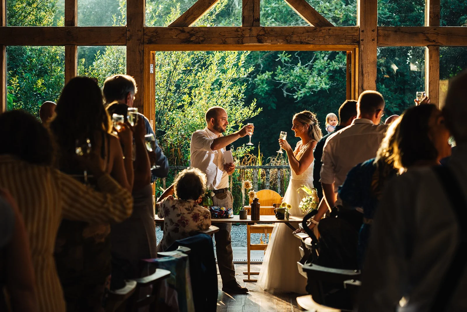 A wedding reception with a bride and groom making a toast inside a wooden venue, surrounded by guests, with sunlight streaming through open doors revealing greenery outside.
