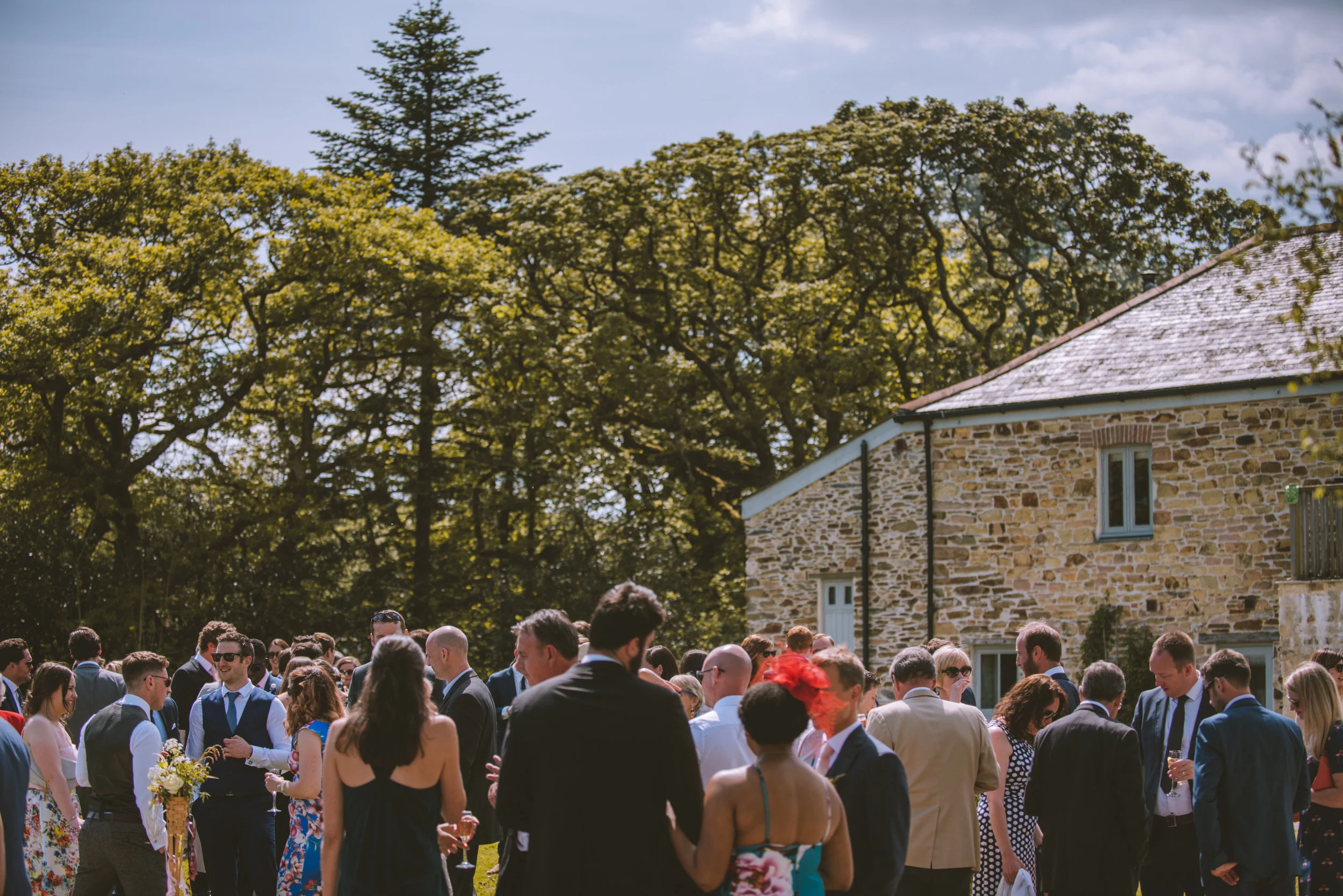 A group of people in formal attire gathered outdoors at a social event, with trees and a stone building in the background.