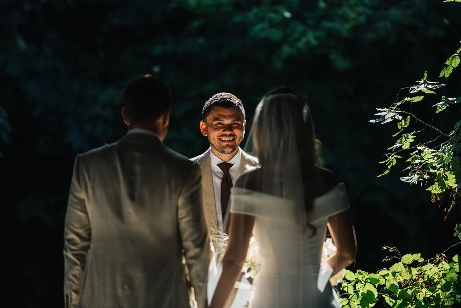 A wedding ceremony outdoors with a bride and groom facing a minister, surrounded by dark greenery.