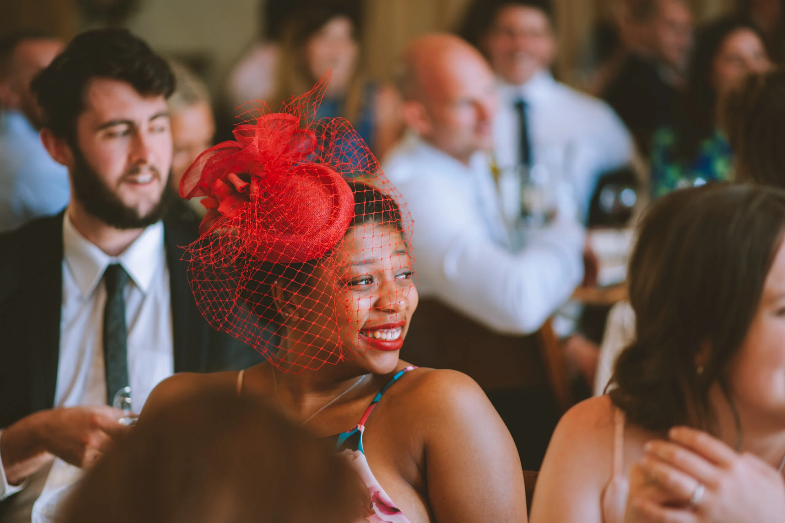 A woman wearing a vibrant red fascinator hat with netting, attending an event or celebration with other people around her.