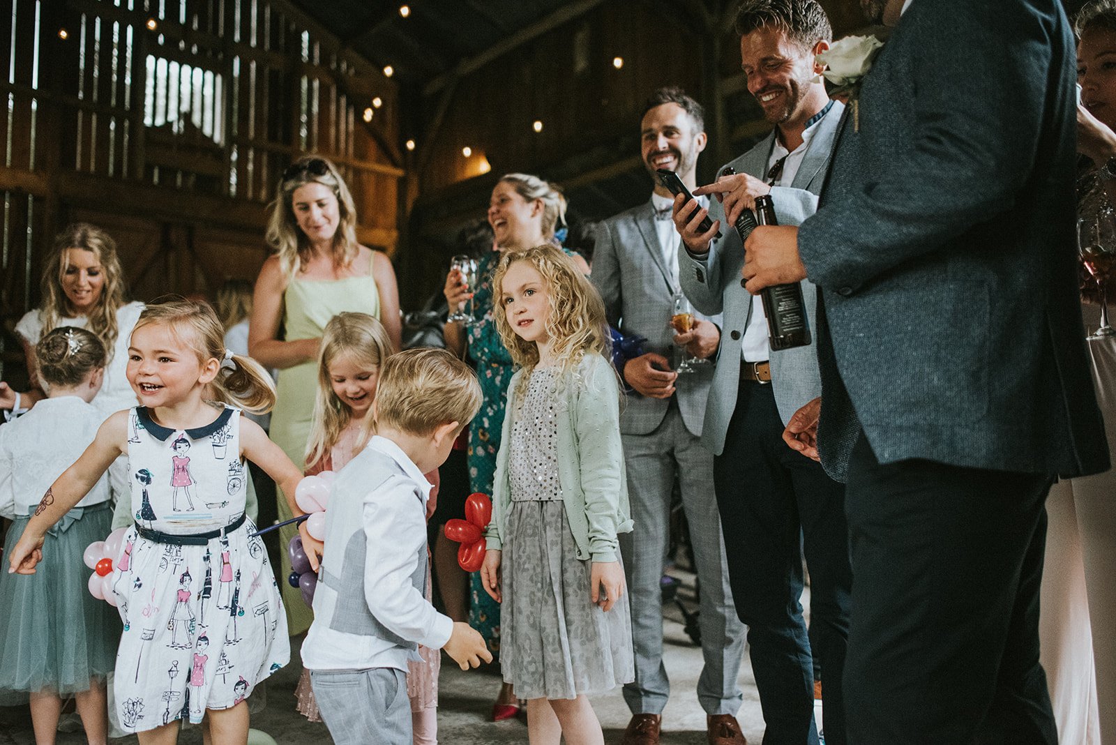 Children and adults at a celebration in a rustic barn, with children playing with balloons and adults smiling and chatting.