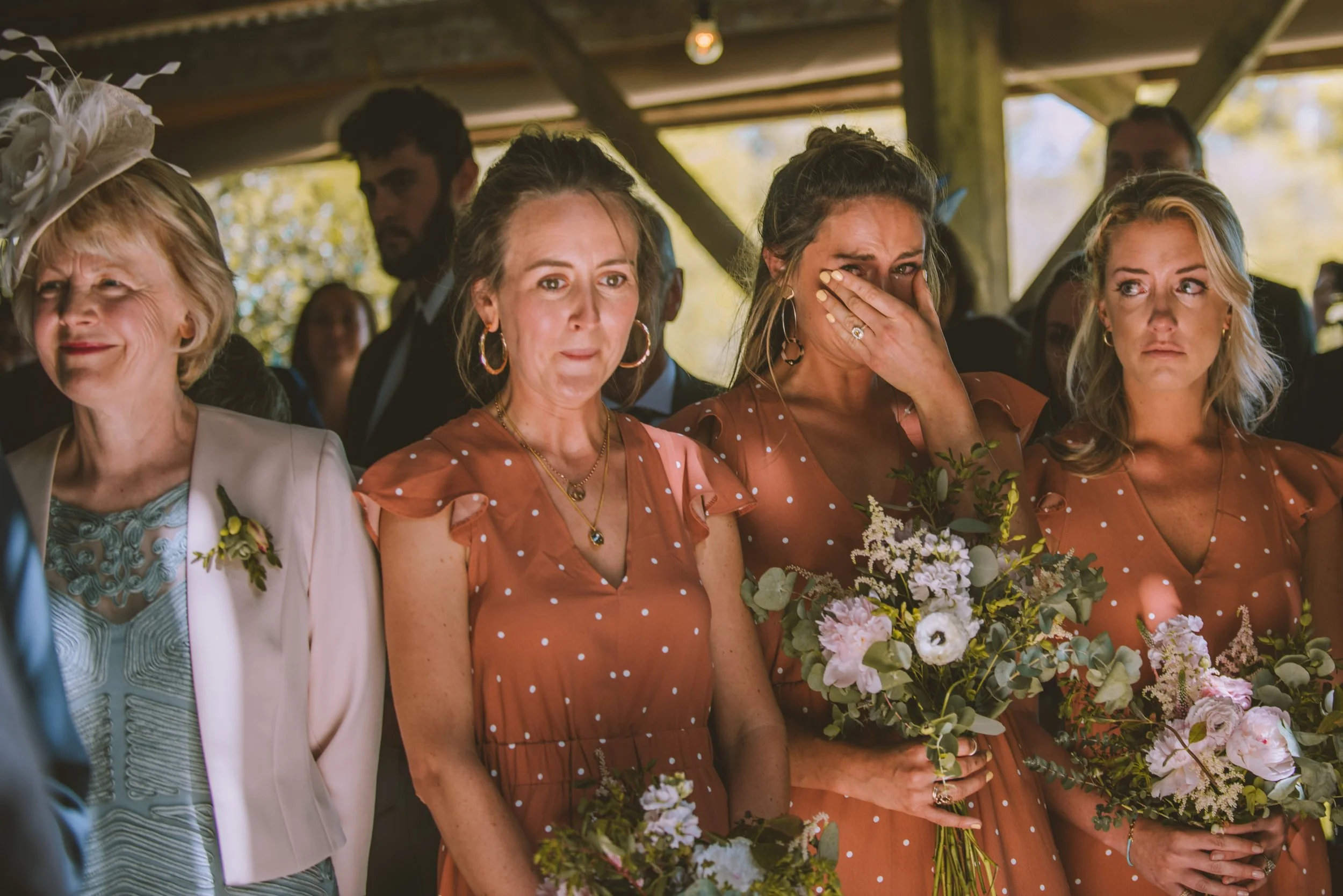 Emotional event with women holding flowers, some tearing up and crying, gathered in a rustic outdoor setting under a wooden structure.