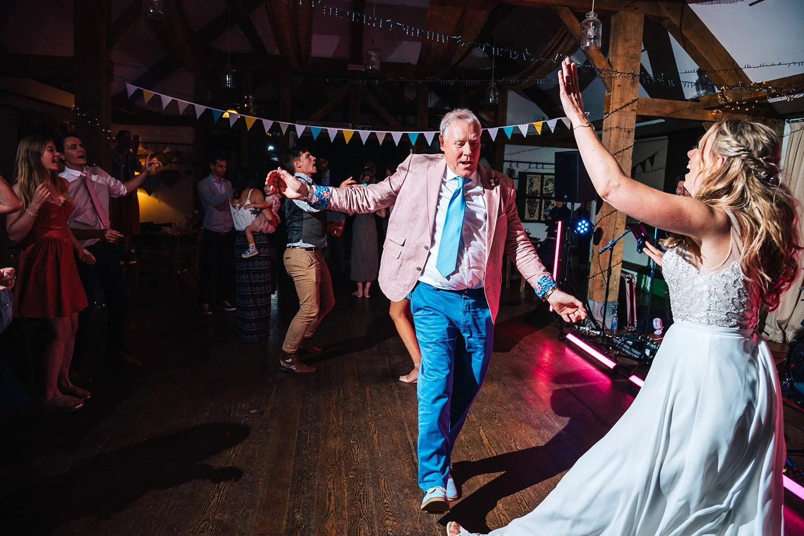 People dancing and celebrating at a wedding reception in a decorated venue with bunting and string lights.