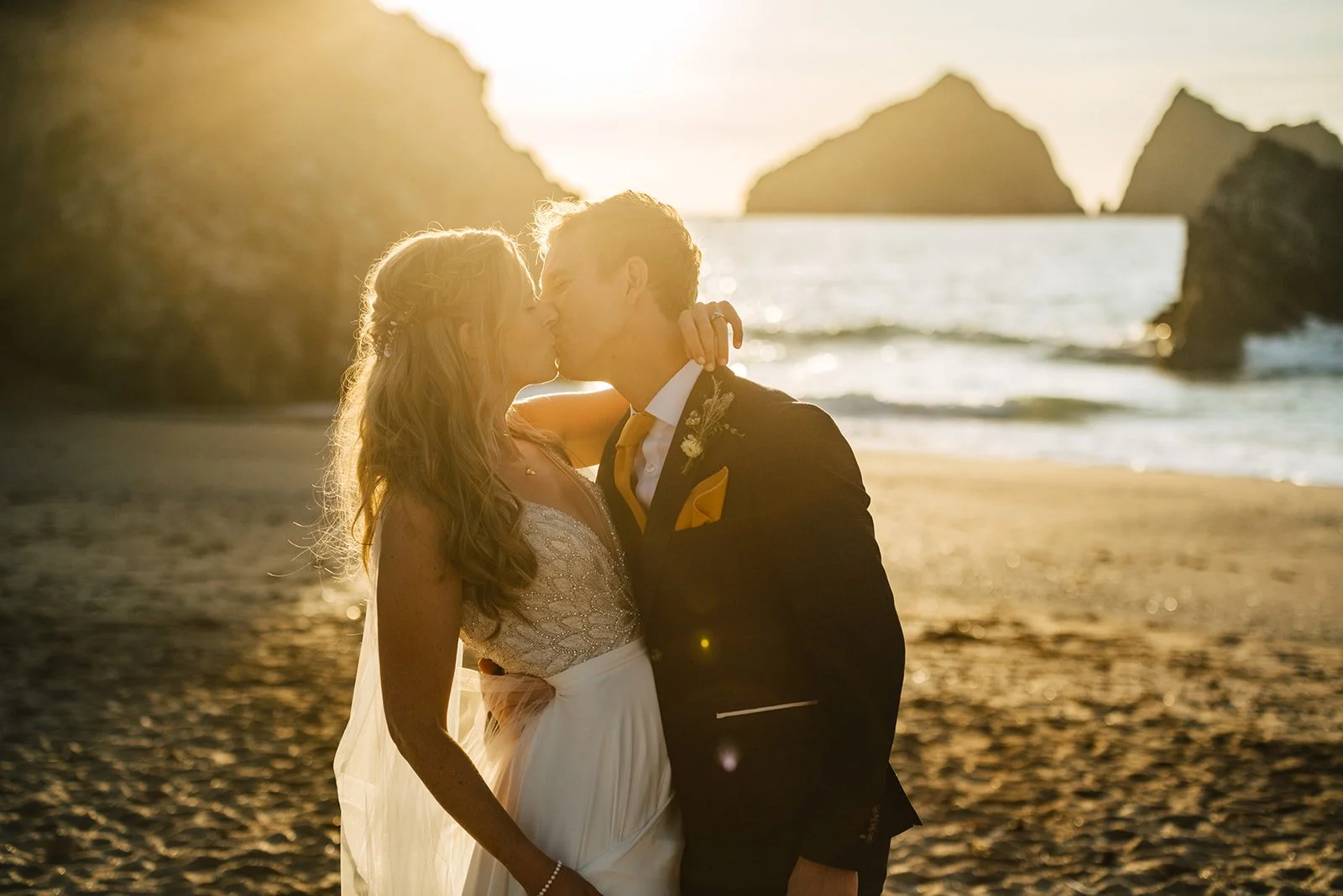 A couple kissing on the beach during sunset, dressed in wedding attire.