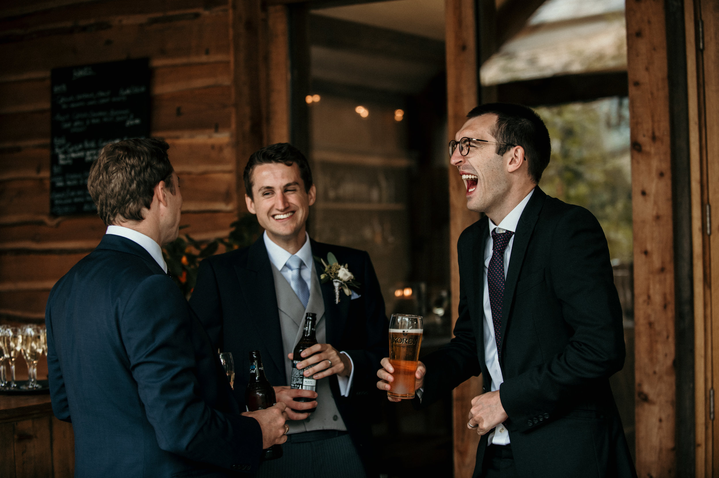 Three men dressed in suits are laughing and enjoying drinks at a social gathering in a rustic wooden interior.