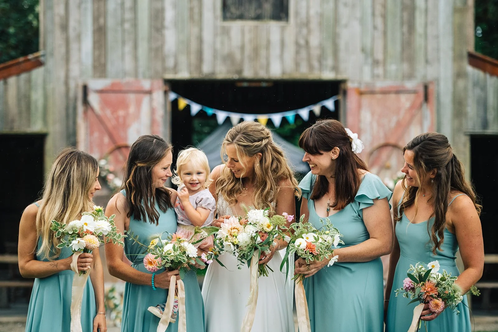 A bride holding a bouquet surrounded by five women in teal dresses and a young girl in a light dress, smiling and laughing in front of a rustic barn with bunting decorations.