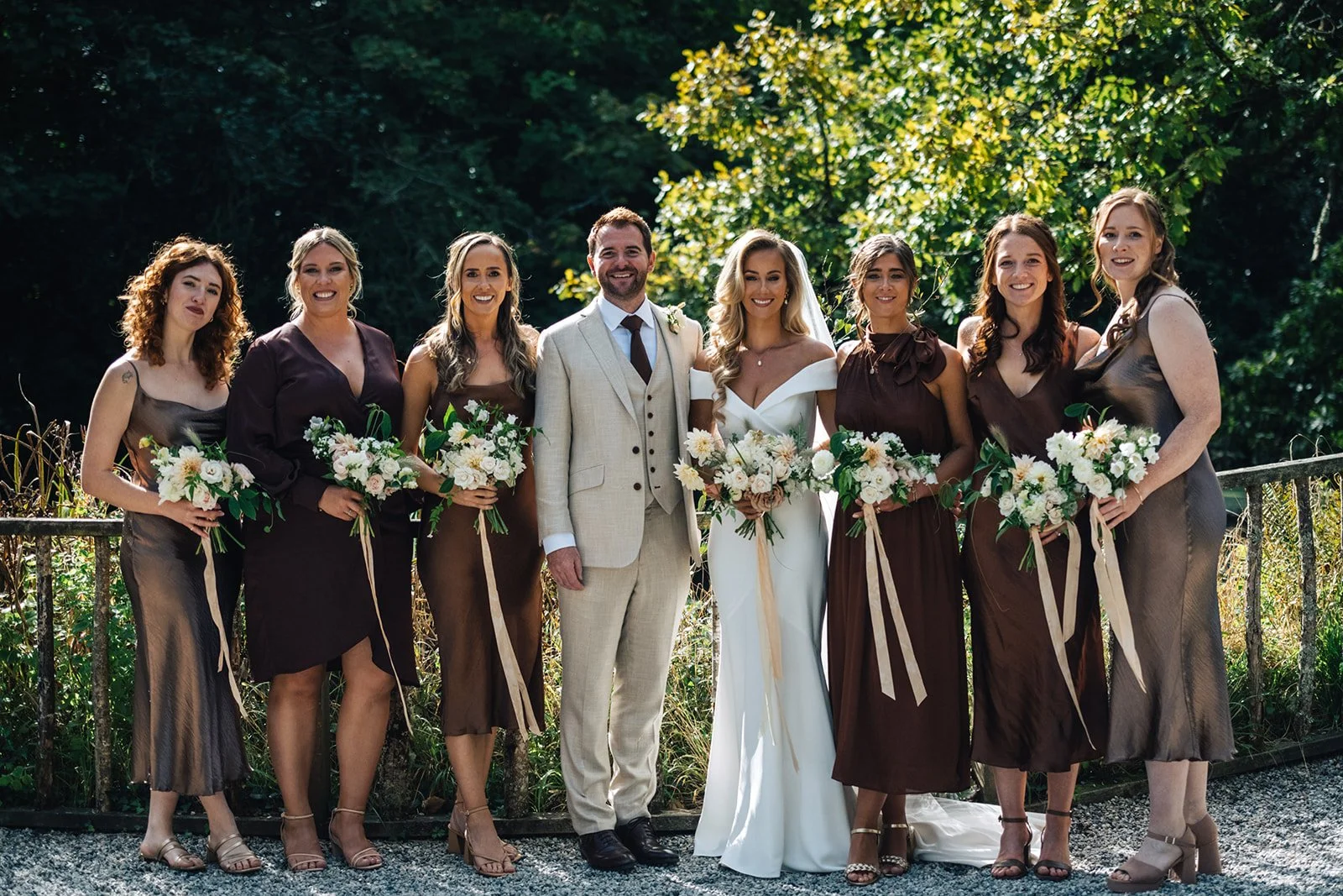 A wedding party of eight standing outdoors, including a bride in a white dress, a groom in a beige suit, and six bridesmaids in brown dresses, all holding floral bouquets.