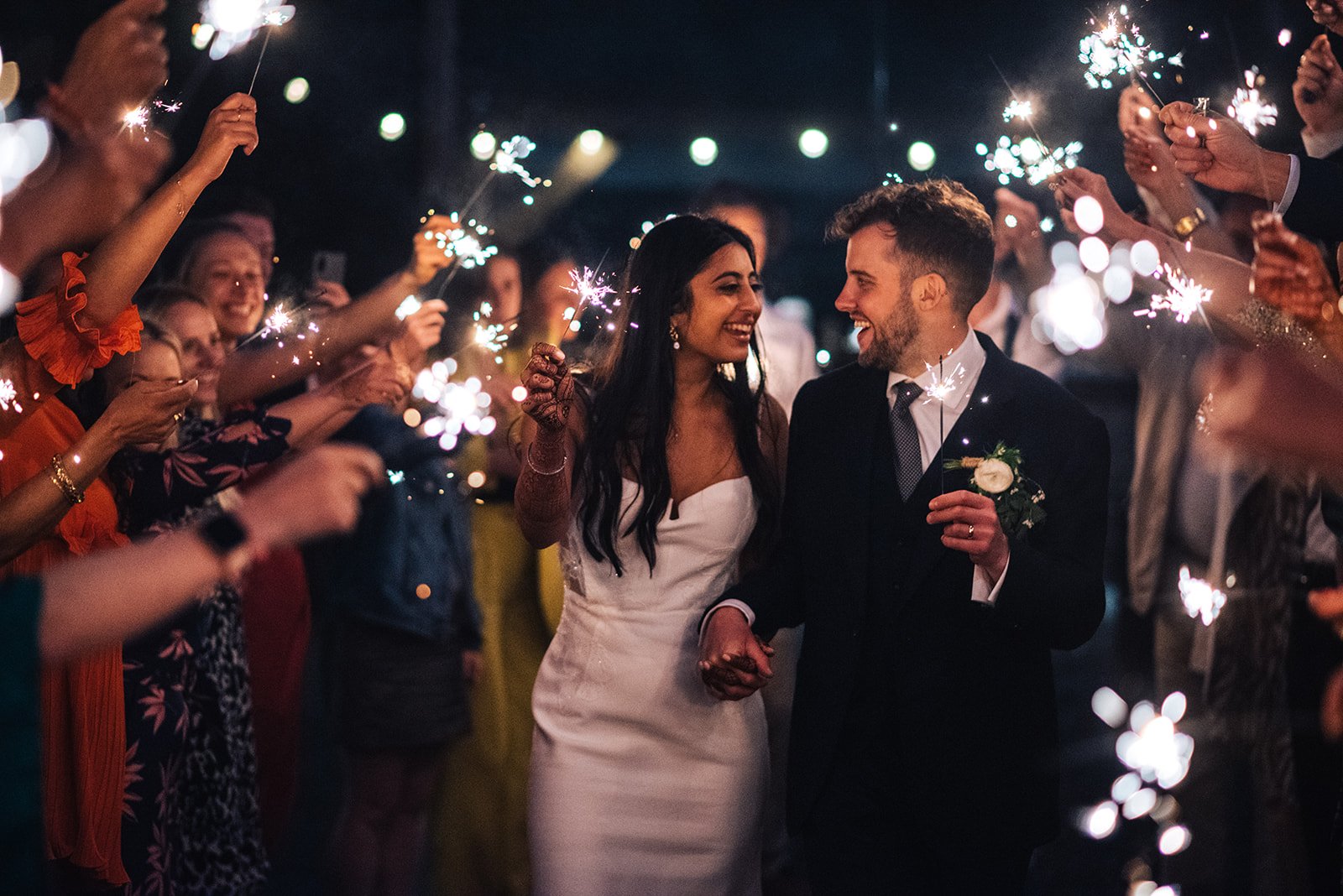 A newlywed couple holding hands and smiling at each other while surrounded by celebrating guests holding sparklers at night.