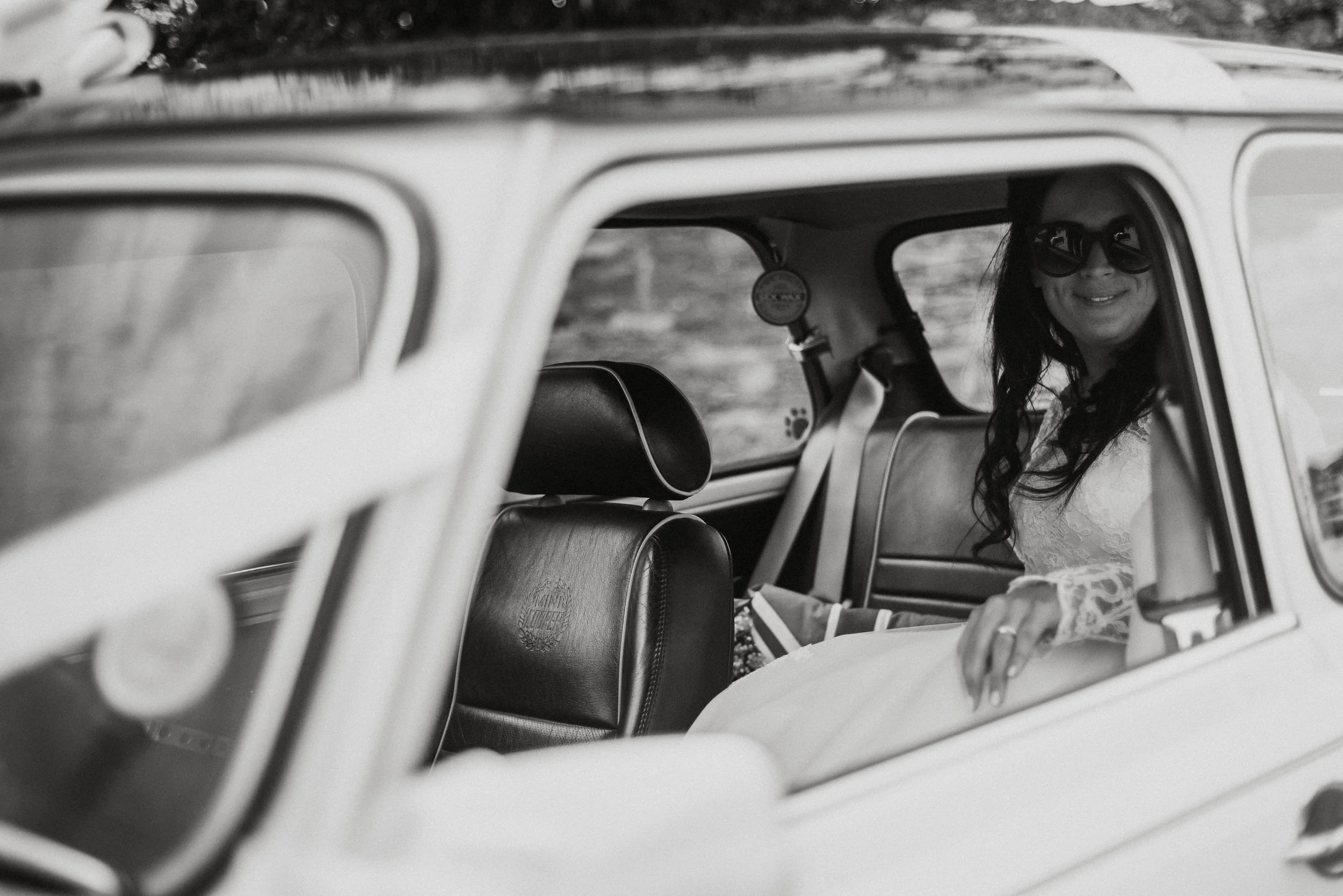A woman with long dark hair, wearing sunglasses and a white lace dress, sitting in the back seat of a vintage car, smiling at the camera.