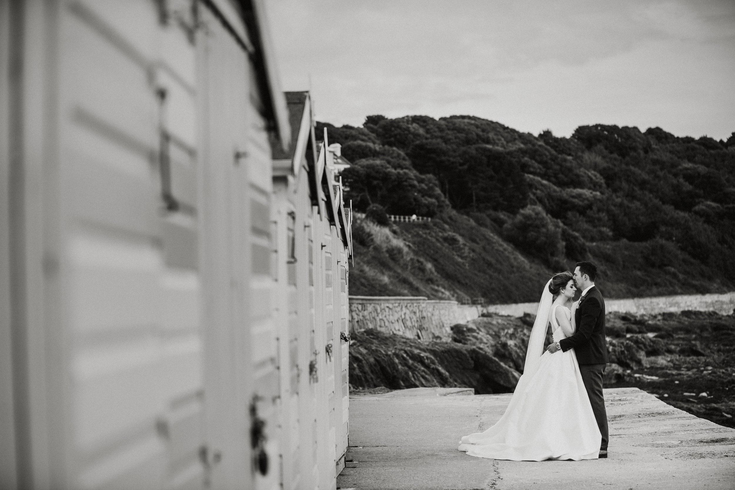 Black and white photo of a bride and groom kissing on a concrete pathway by the ocean, with a hillside covered in trees in the background.