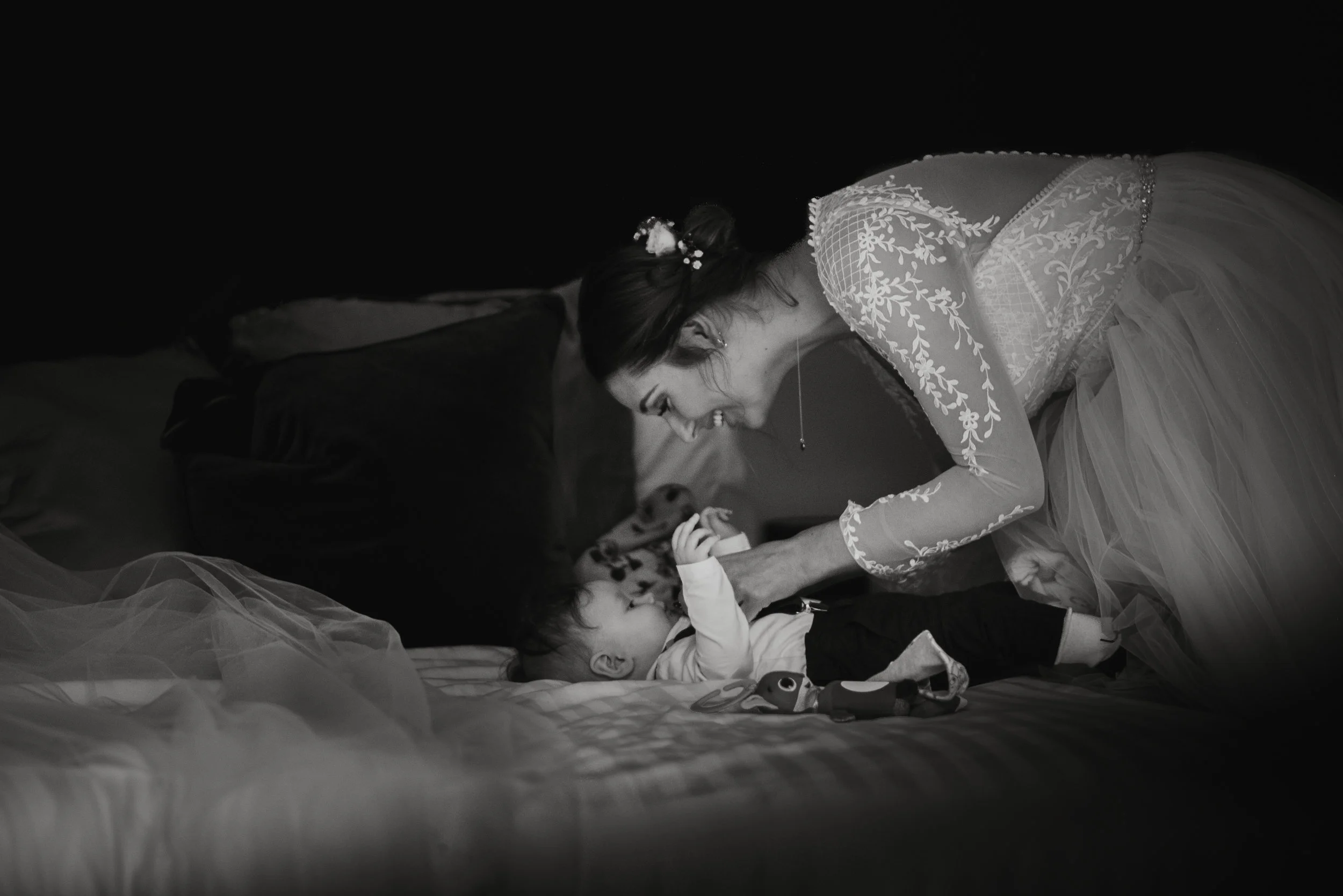 A woman in a wedding dress smiling and playing with a baby on a bed in a dimly lit room.