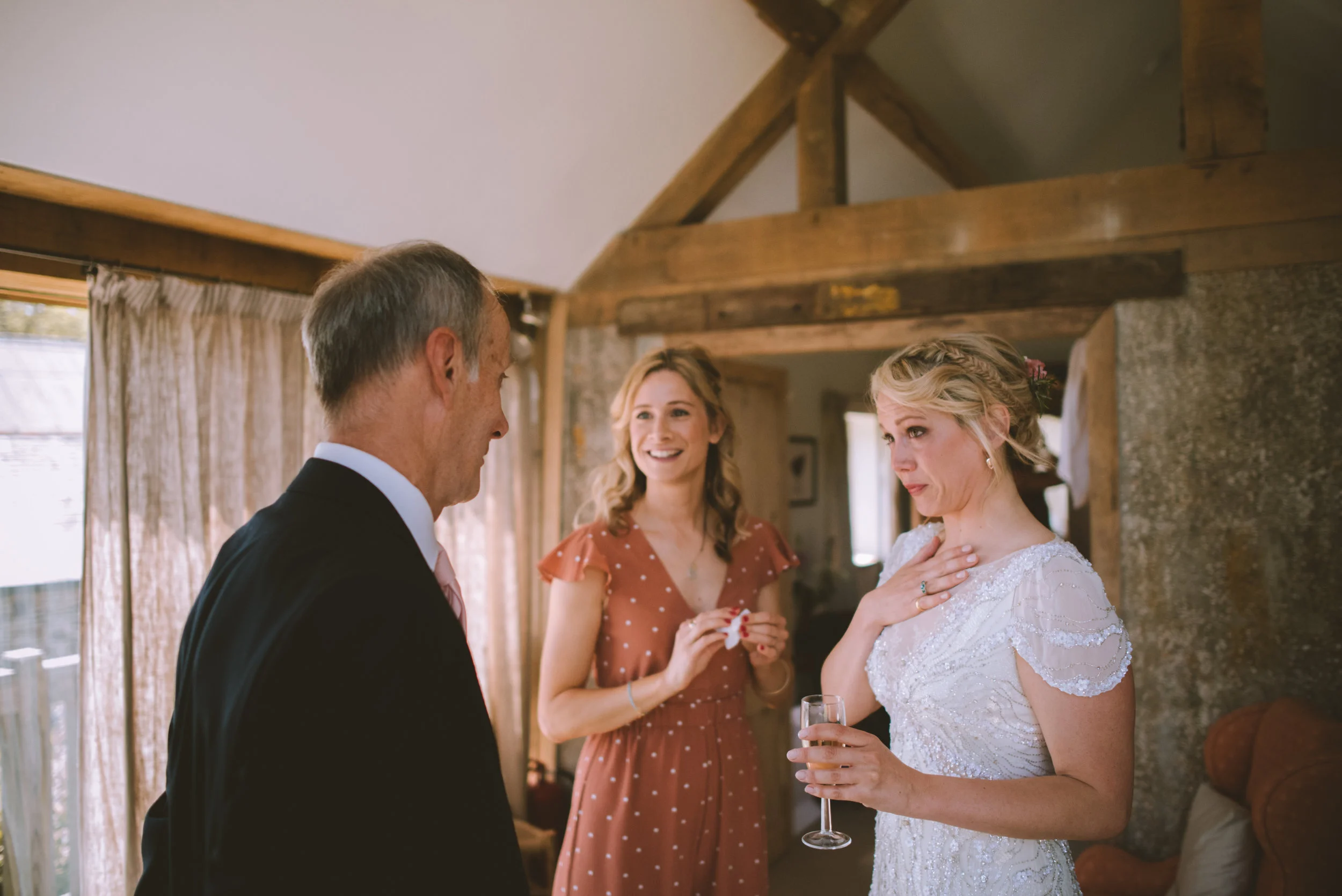 A bride in a white wedding dress with a microphone in her hand, a man in a suit, and a woman in a rust-colored dress with white polka dots inside a rustic venue with wooden beams.