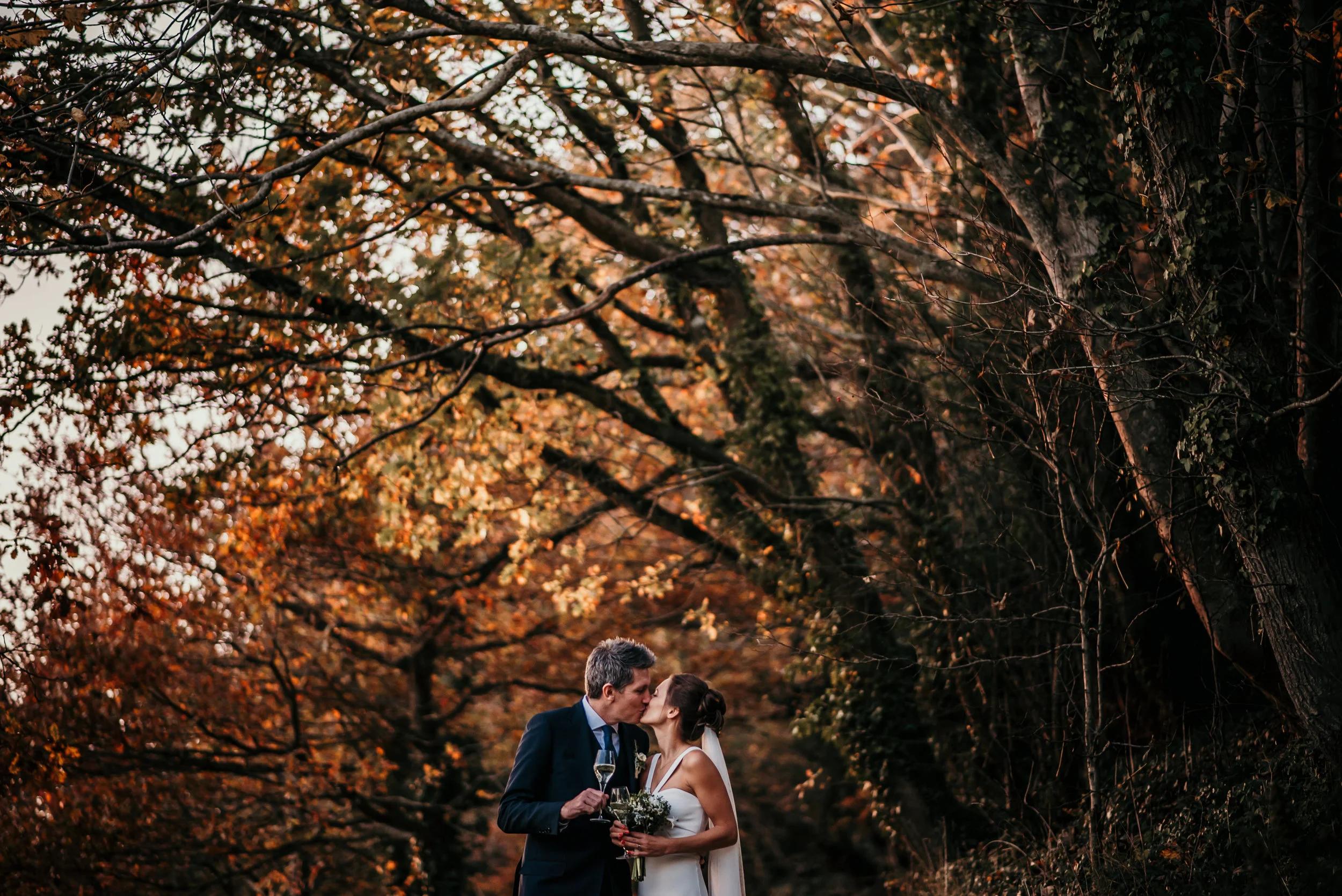 A bride and groom kissing outdoors during sunset, surrounded by autumn-colored trees, with the bride holding a bouquet and the groom holding a glass of champagne.