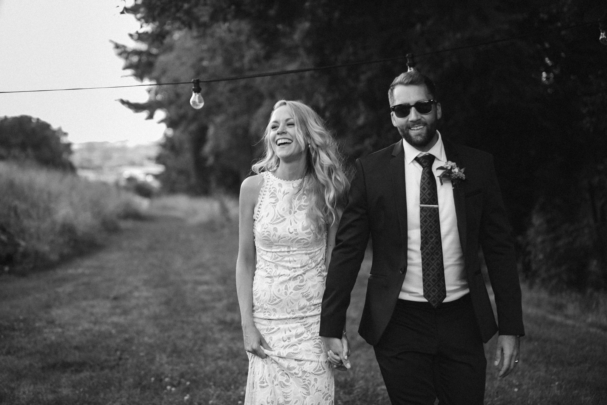 A black and white photo of a smiling woman in a lace dress holding hands with a man in a suit with sunglasses, walking outdoors on a grassy path with trees in the background.