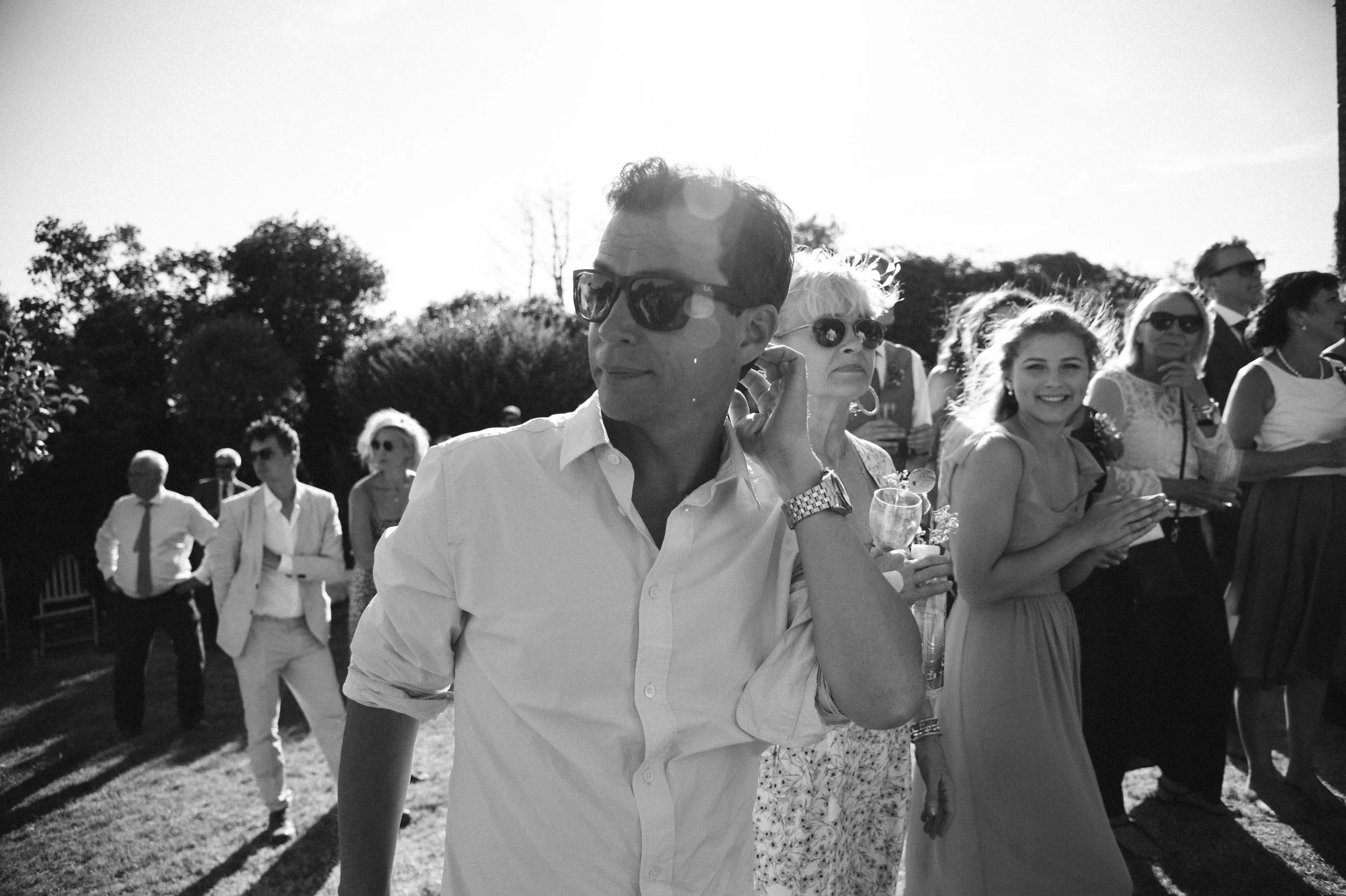 Group of people at an outdoor event, dressed in formal attire, some holding drinks, with trees and clear sky in the background.