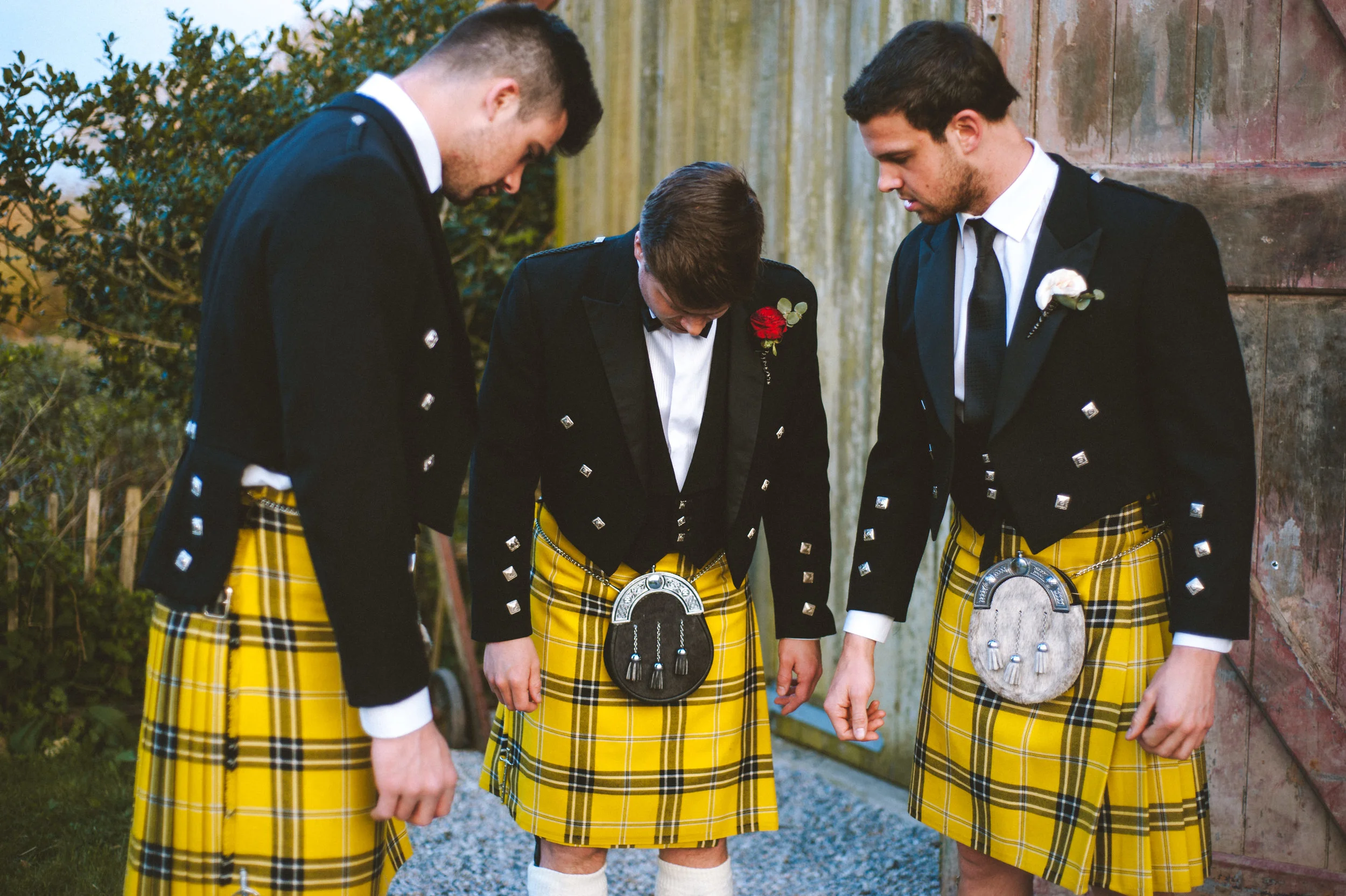 Four men in traditional Scottish kilts and black jackets standing outdoors, bowing their heads in a moment of reflection or prayer.
