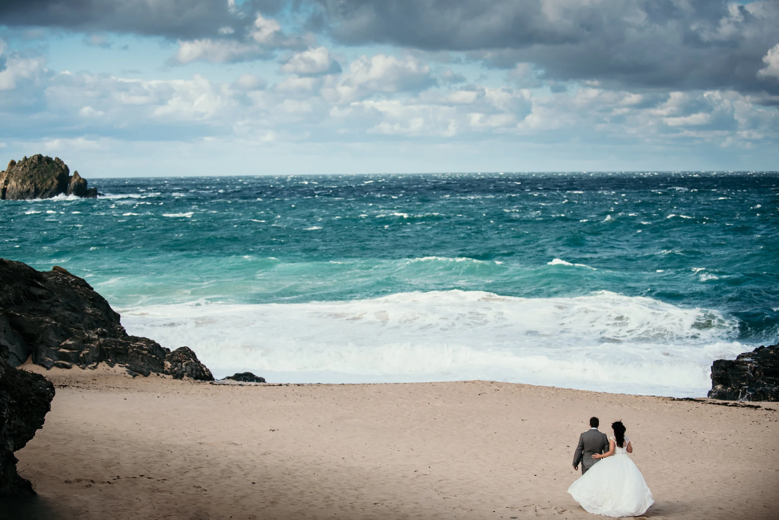 A couple in wedding attire walking on a sandy beach near the ocean with rocky formations, blue sky, and clouds.
