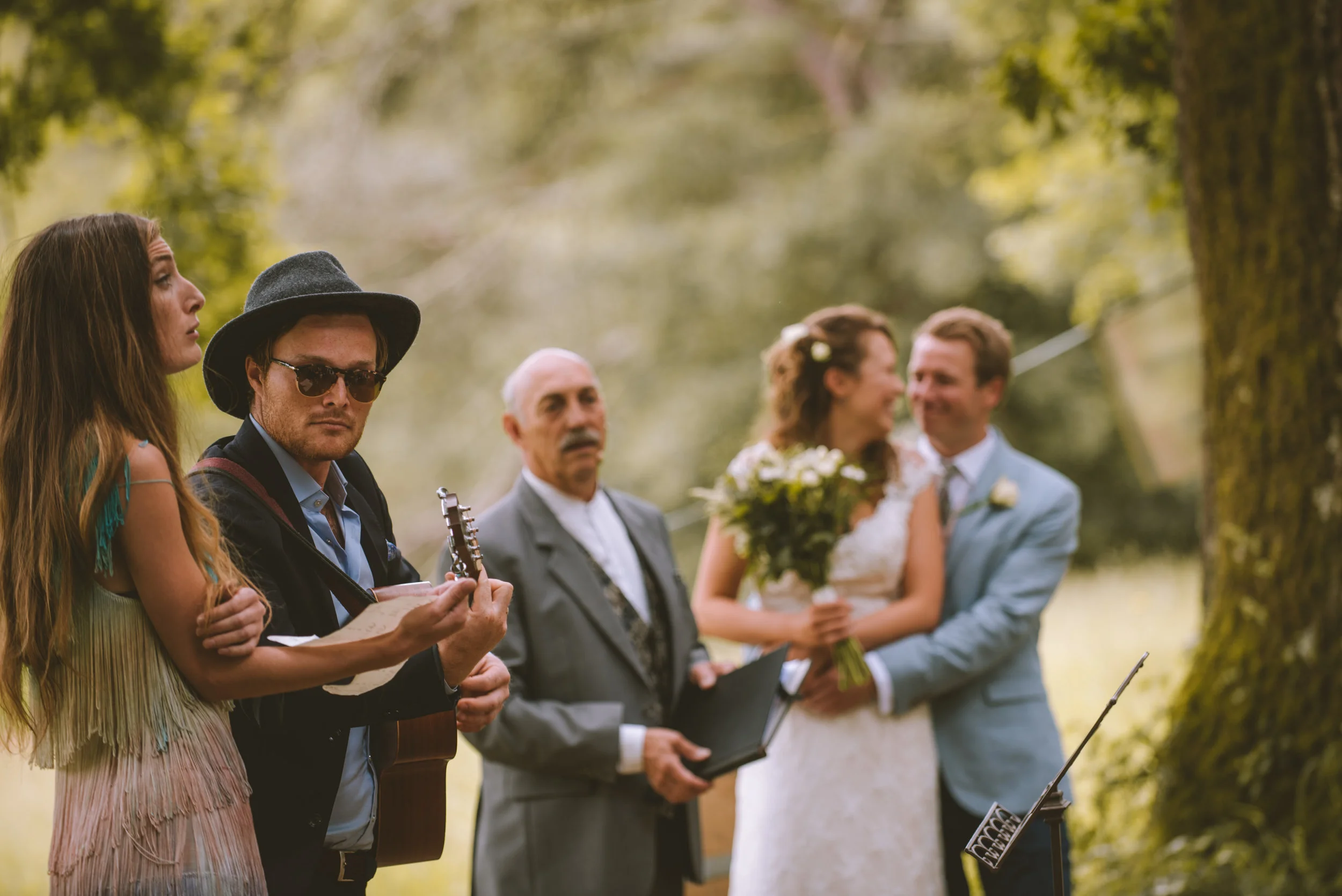 A wedding ceremony outdoors with the bride and groom smiling, holding each other, near a large tree, with an officiant and musician in the foreground, and a woman and man standing nearby.