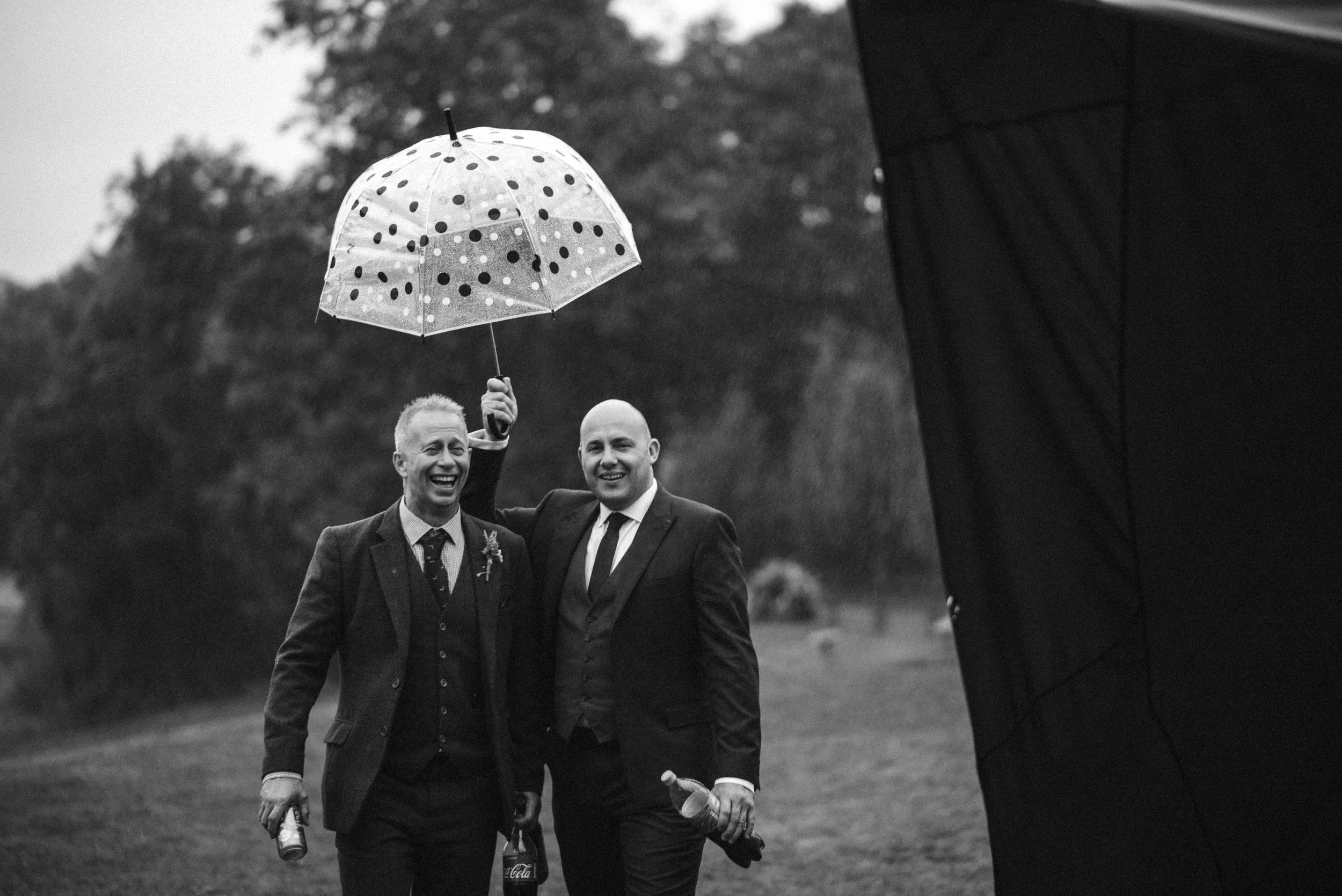 Two men in suits standing outdoors in the rain, smiling, with one holding a polka-dotted umbrella, and both holding drinks, with trees in the background and a large umbrella or reflector on the right.