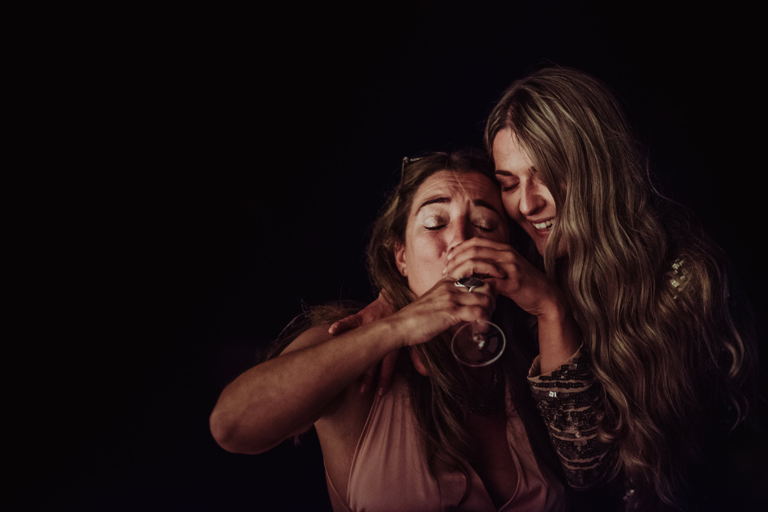 Two women sharing a joyful moment, one holding a wine glass, in a dark setting.