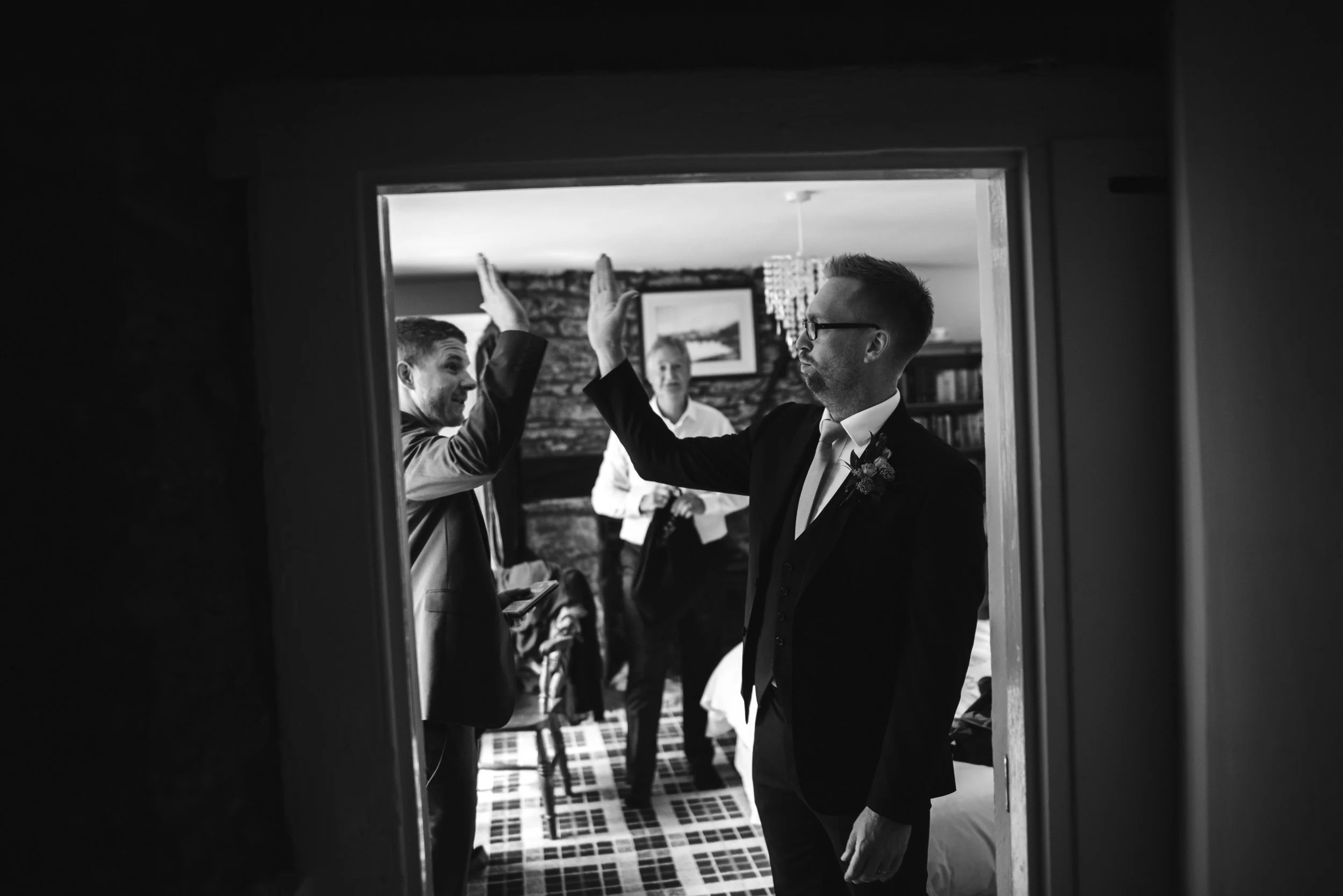 Black and white photo of a groom and groomsman giving each other a high five inside a room with other people in the background.