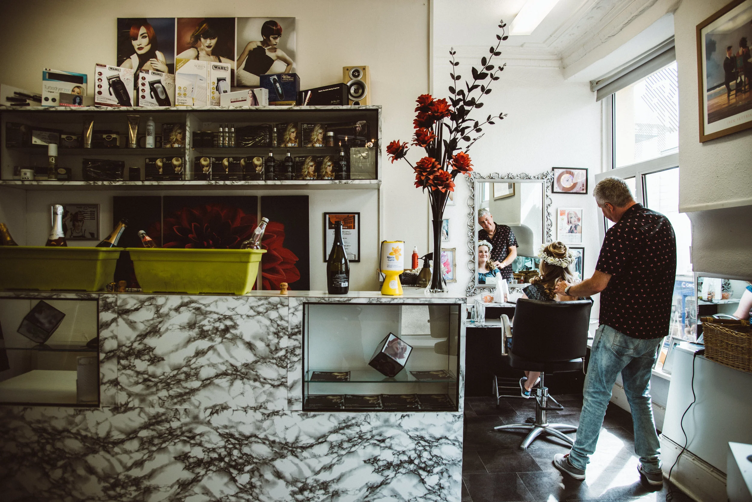 A hair salon with a stylist working on a client's hair near a large mirror, in a space decorated with flowers and photographs, with shelves of beauty products and a marble counter.