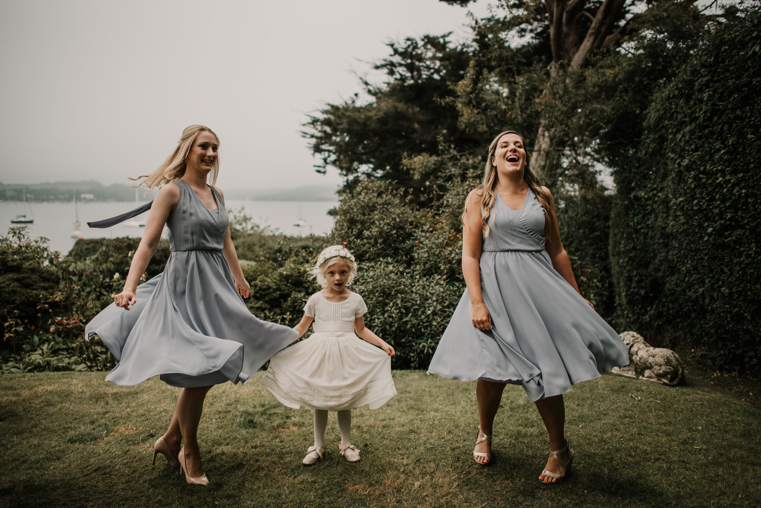 Three women and a young girl enjoying a dance outdoors near a lake, with trees and boats in the background.