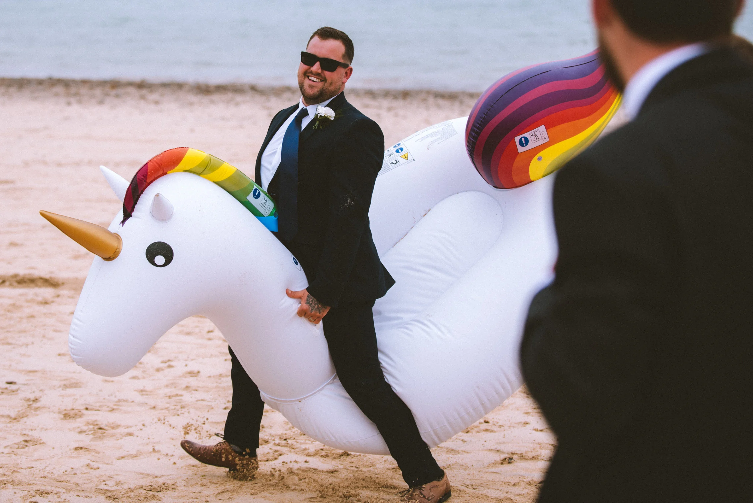 A man in a suit and sunglasses riding on an inflatable unicorn. Another person in a suit is visible in the foreground.