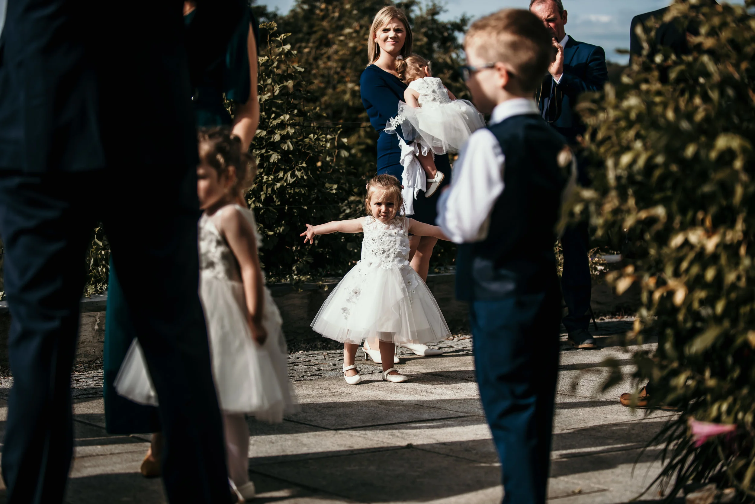 Children in white dresses and suits standing outdoors among adults, with a woman holding a girl in a white dress, in a garden or park setting during daytime.