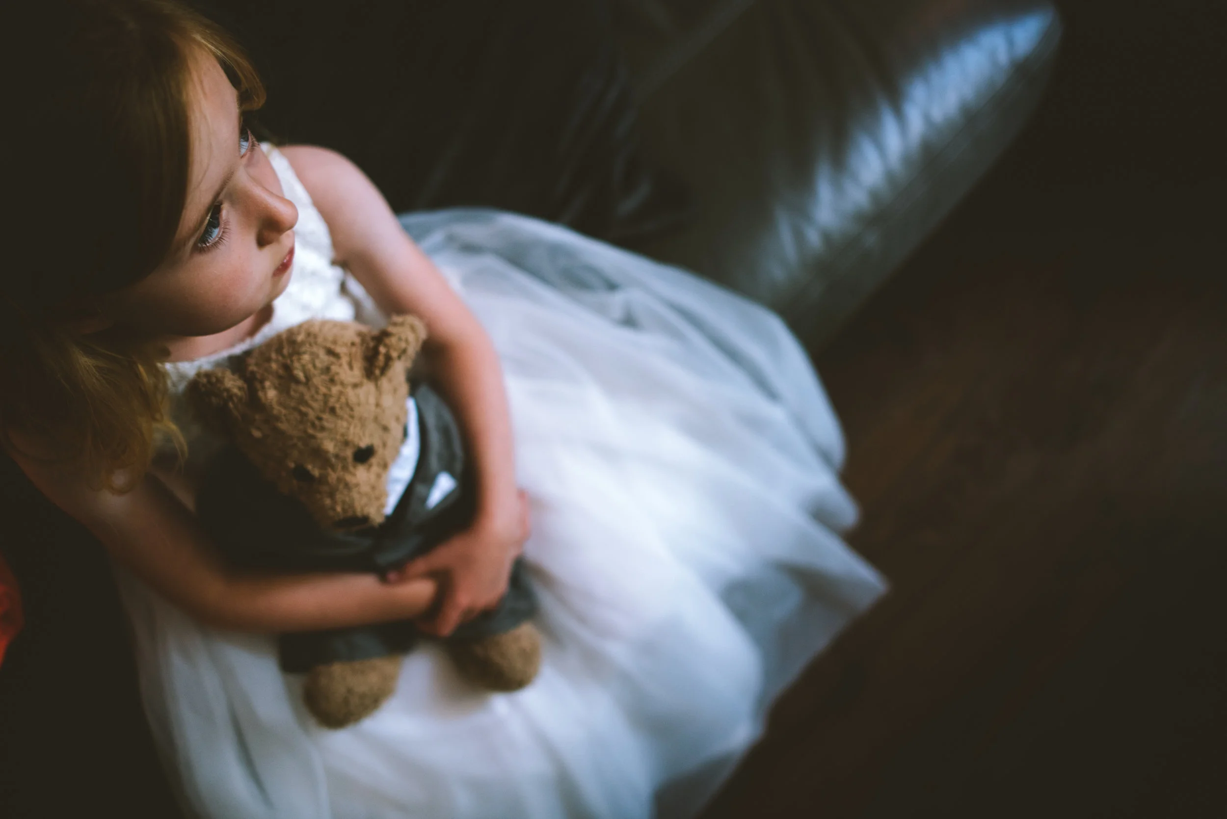 A young girl sitting on a white cushion on a dark brown leather couch, holding a plush teddy bear, with a dark background.