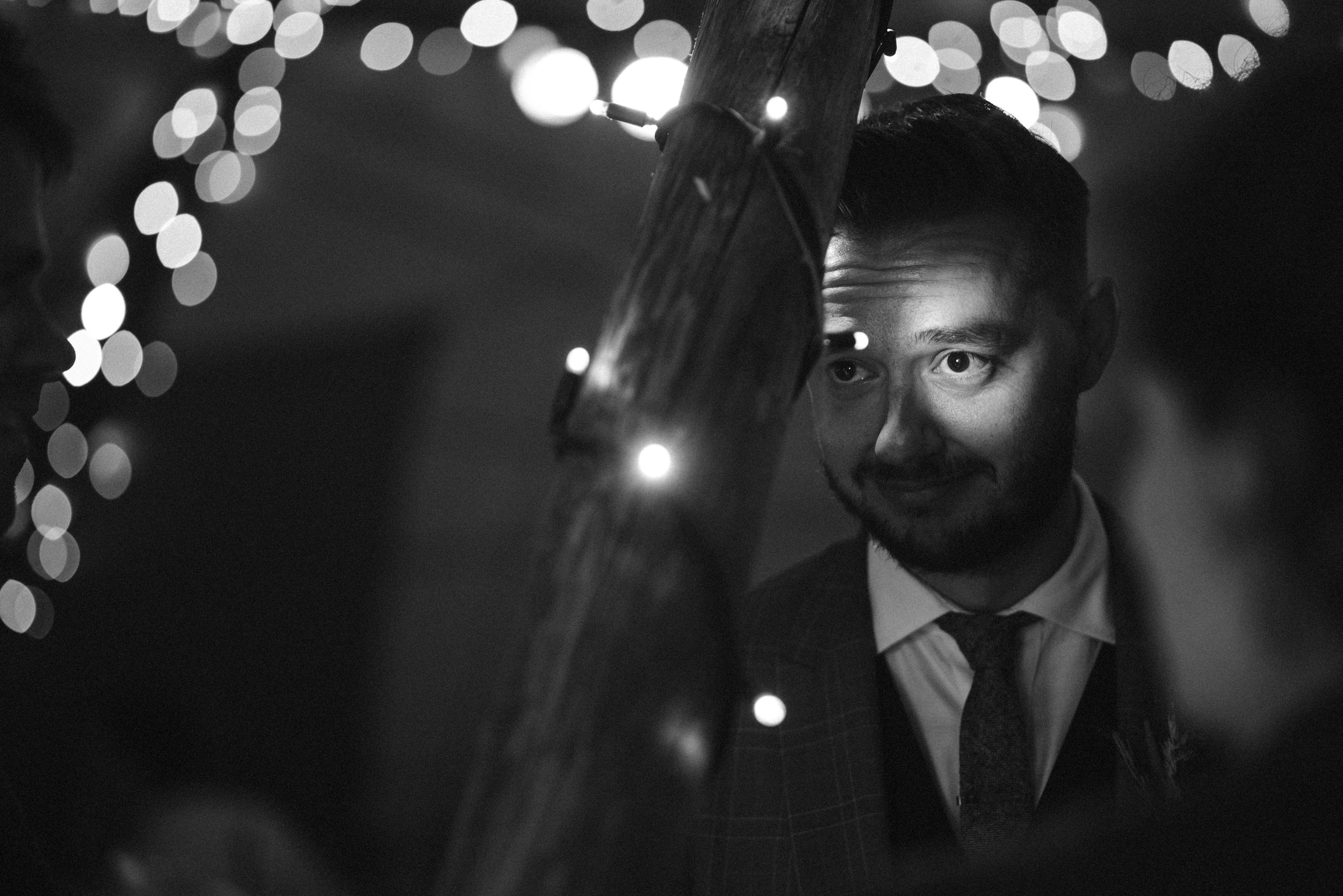 A man in a suit looking at another person at night, with string lights in the background.