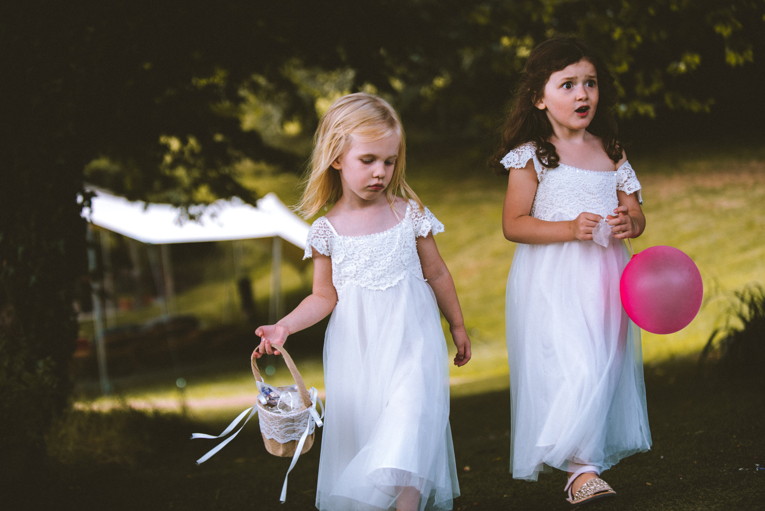 Two young girls in white dresses walking outside in a park-like setting. The girl on the left has blonde hair and is holding a small basket, while the girl on the right has dark hair and is holding a pink balloon. Both girls are wearing lace dresses 