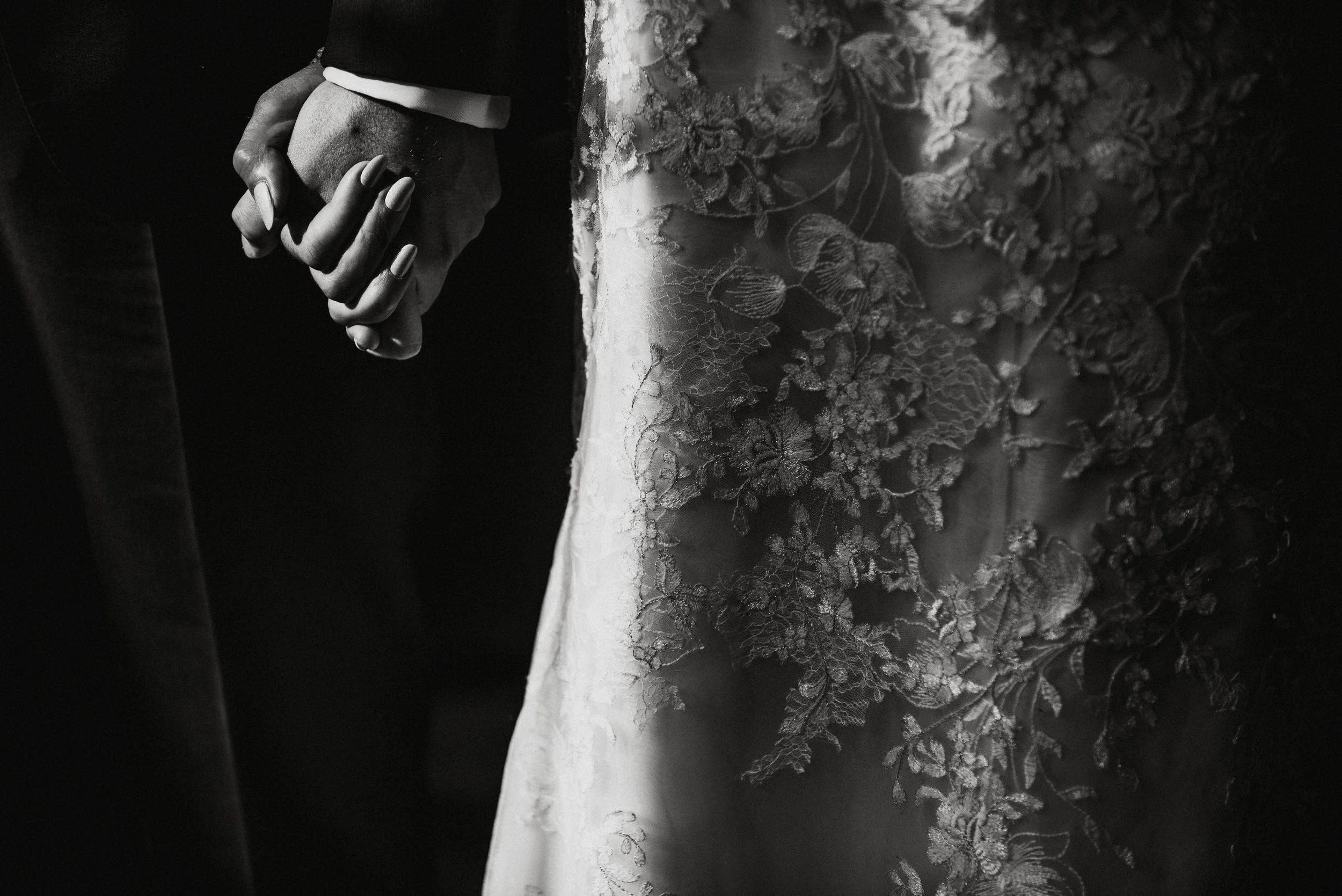 Close-up of a bride and groom holding hands, with the bride wearing a lace wedding dress.