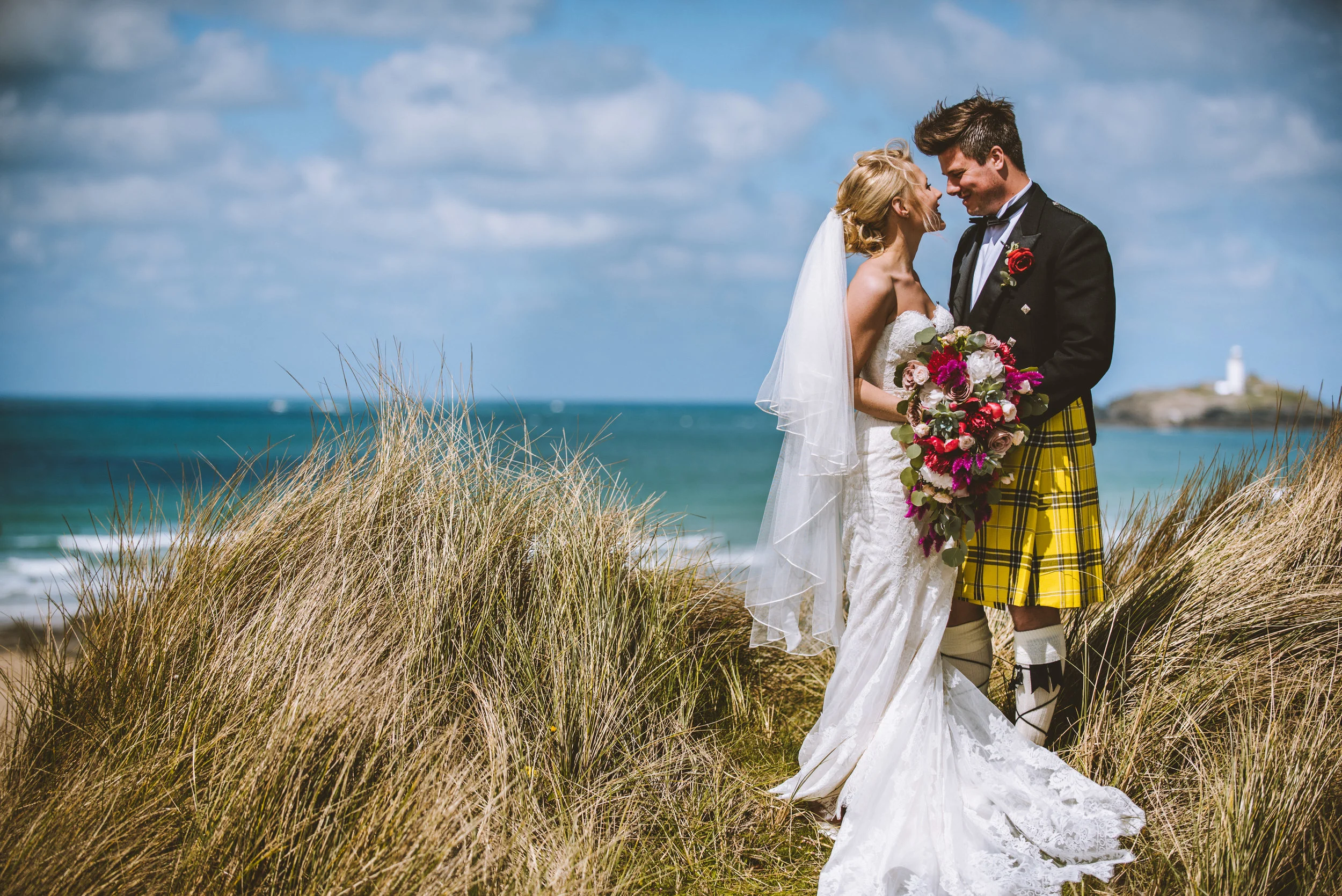 Bride and groom at Gwithian in Cornwall, with the ocean and lighthouse in the background, holding a bouquet of flowers.
