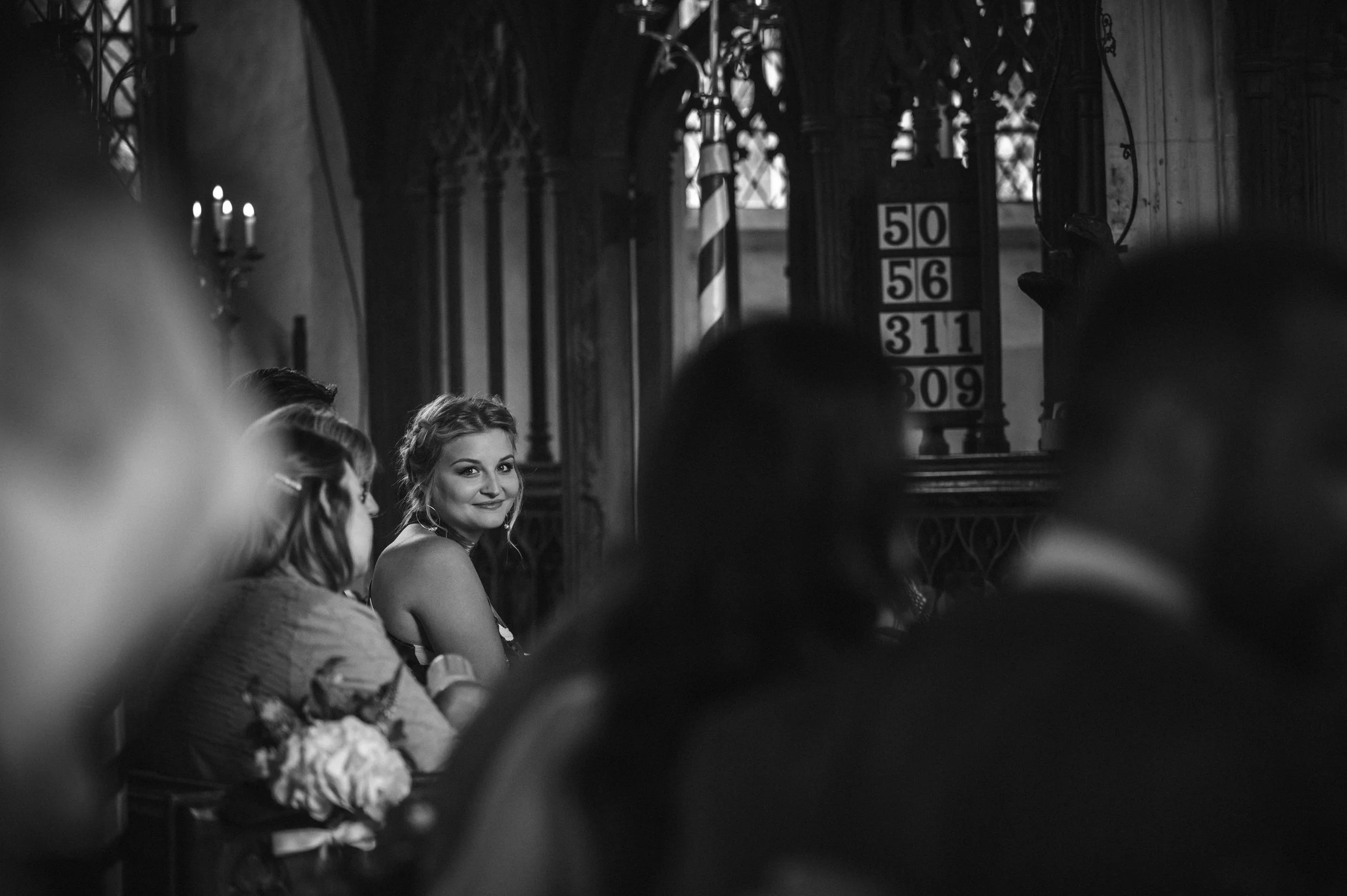 A woman smiling at the camera during a gathering in a church, with other people sitting around her. Church interior with candle holders and hymn boards in the background.