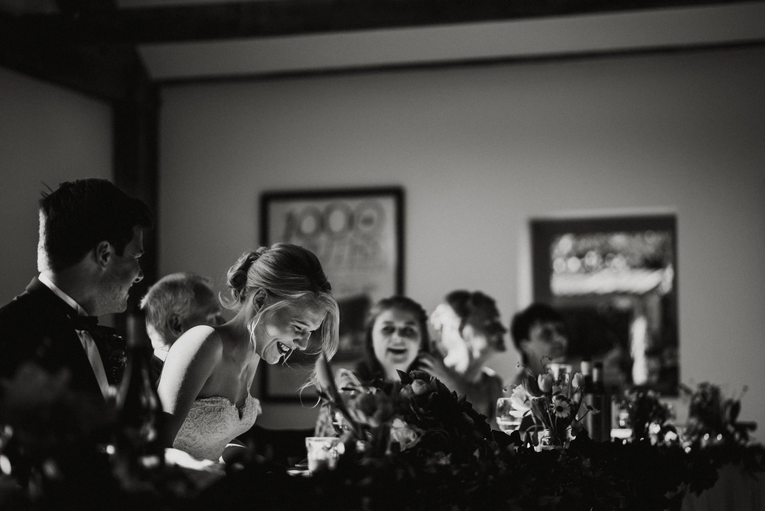 Black and white photo of a wedding reception with a bride laughing and sitting at a table with guests and floral decorations.