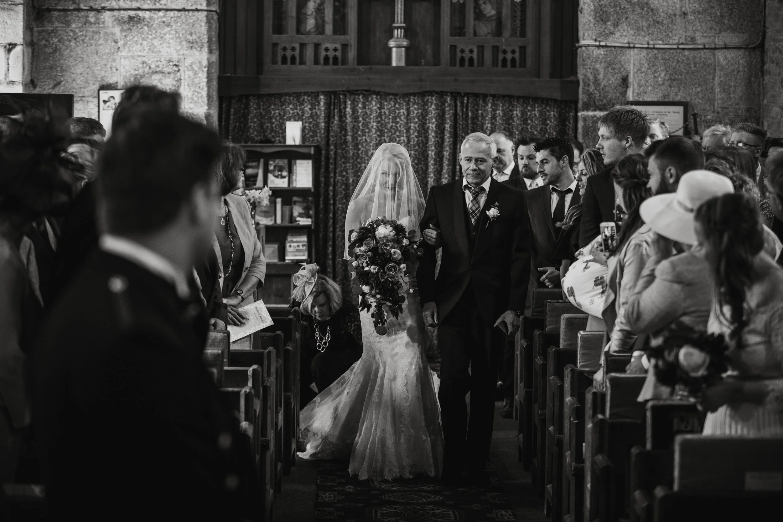 Black and white photo of a bride walking down the aisle with her father at a wedding ceremony inside a church, surrounded by seated guests.