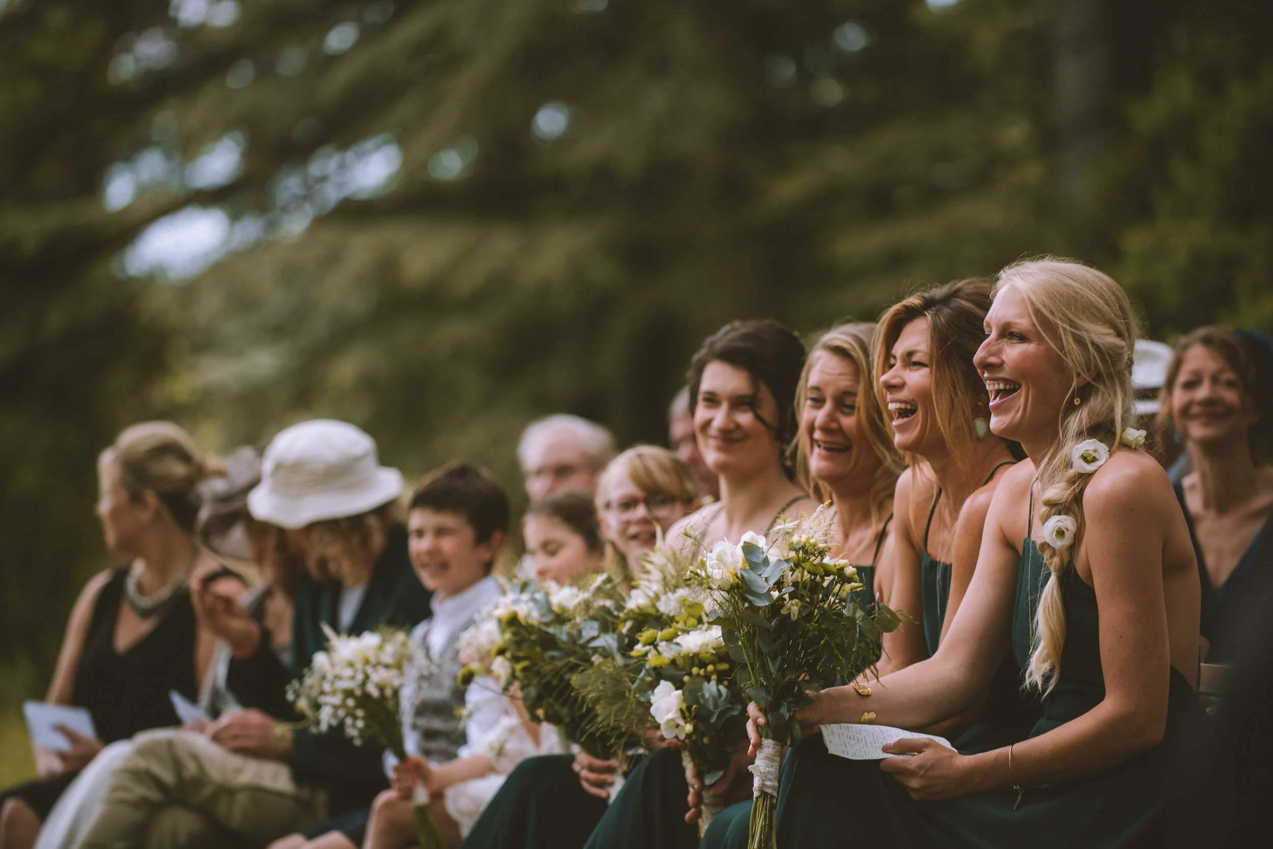A group of people sitting outdoors during a wedding ceremony, smiling and holding bouquets of flowers, with trees in the background.