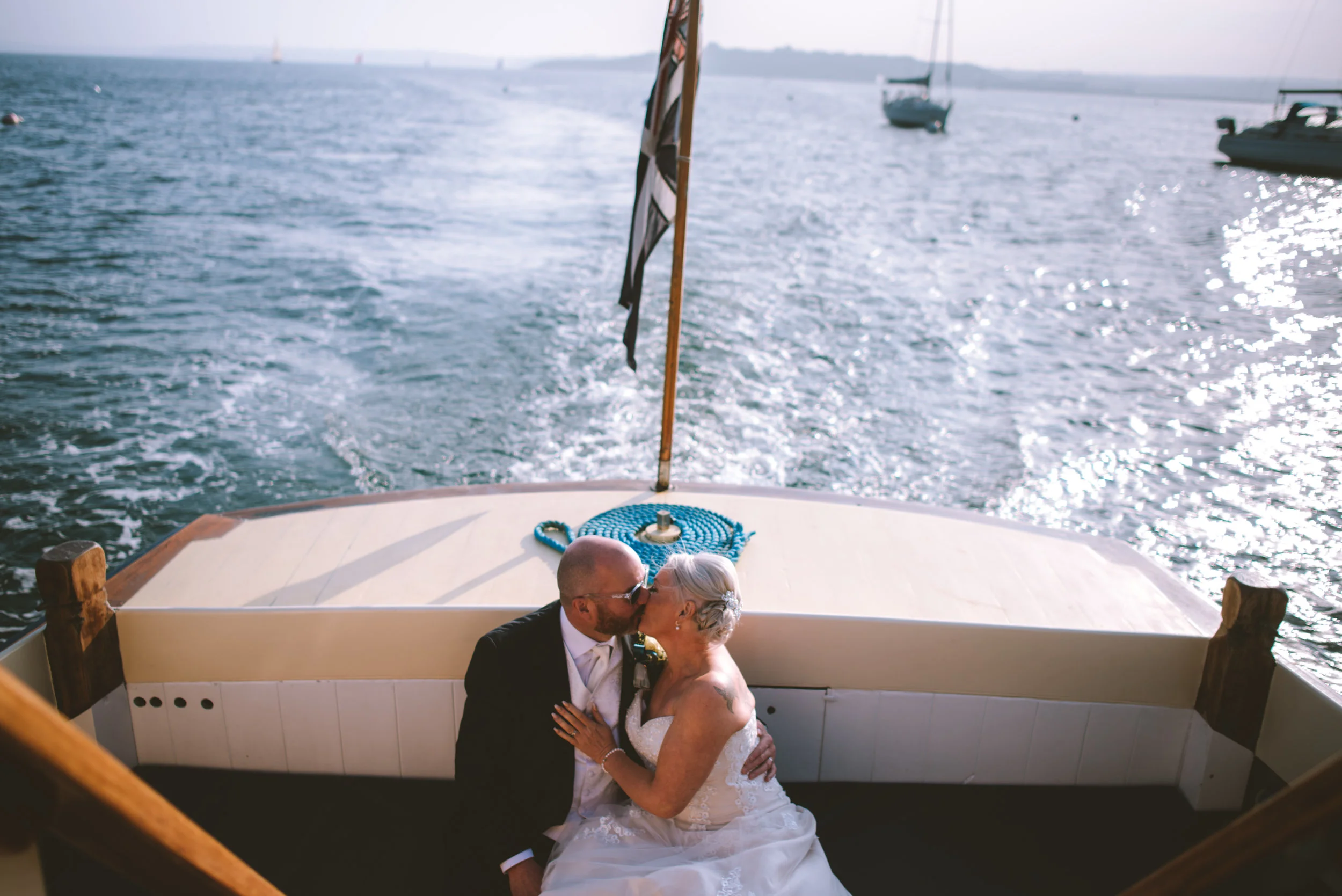 A couple in wedding attire kissing on a boat at sea with other boats in the background during sunset.