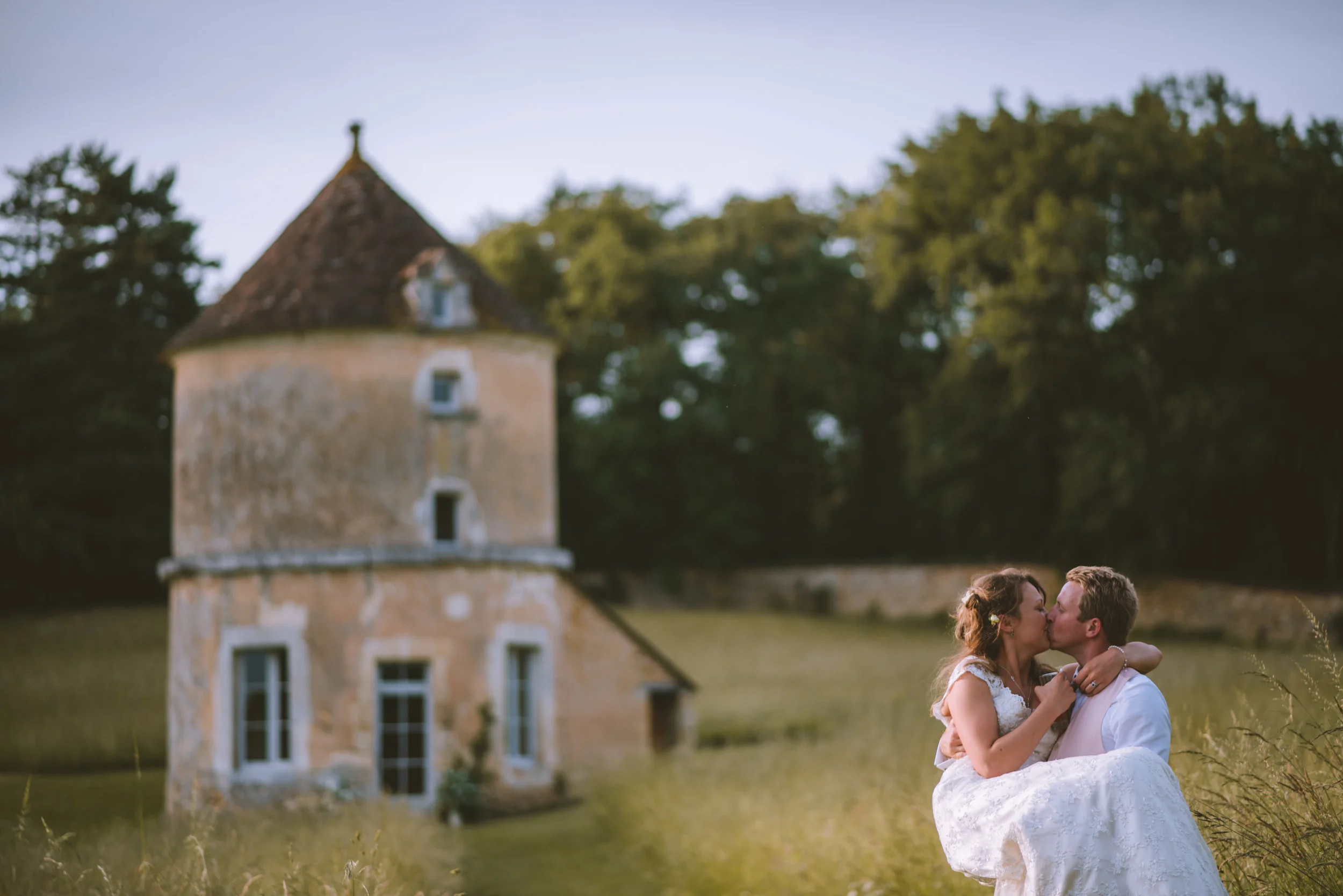 A newlywed couple sharing a kiss outdoors near an old, round stone tower and surrounded by trees during sunset.
