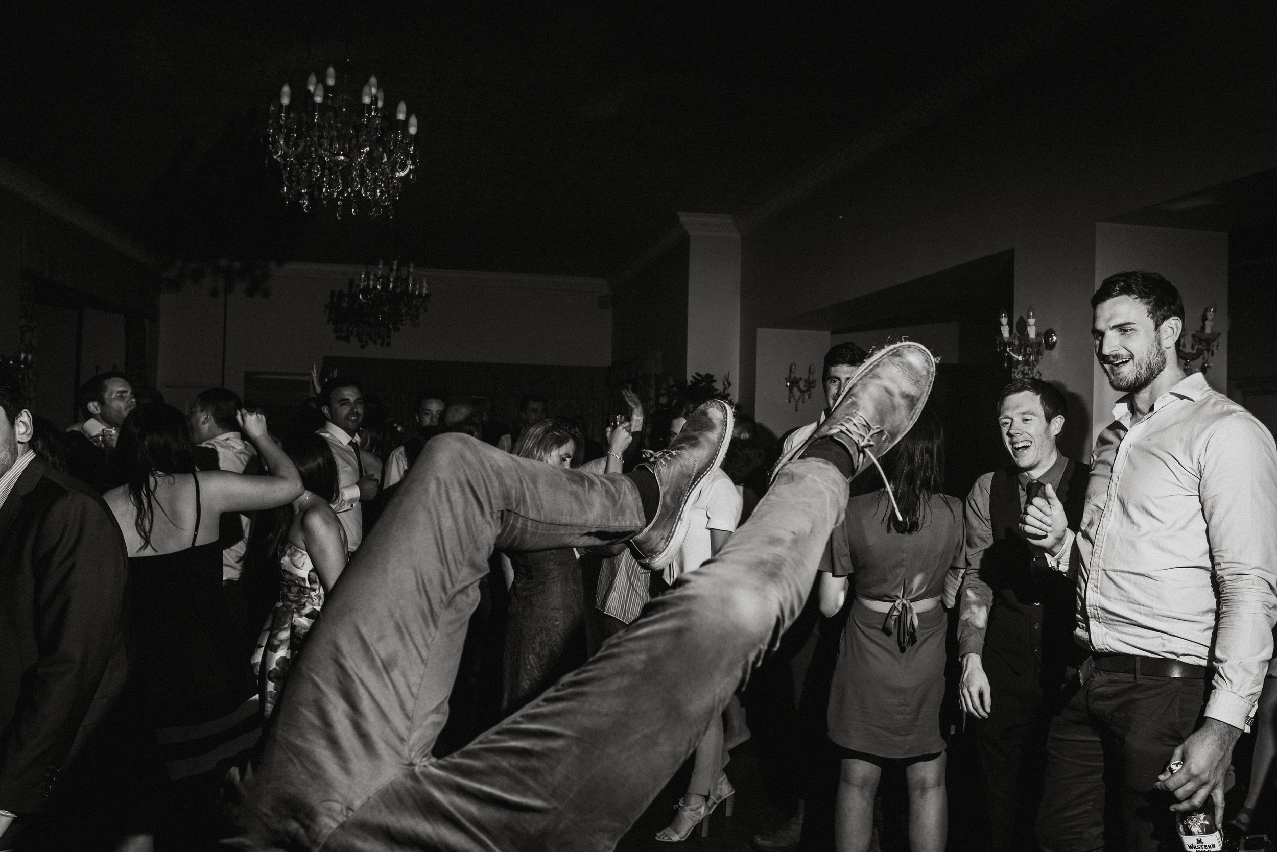 People dancing at a party, with one person’s legs and shoes lifted into the air in the foreground, in a black and white photo.