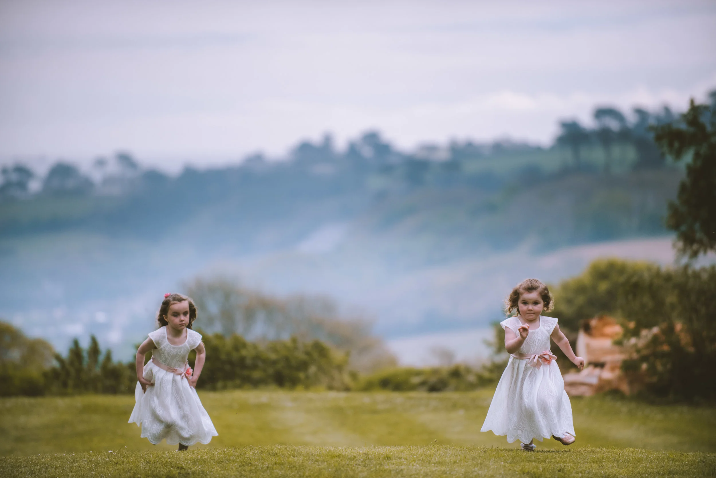 Two young girls in white dresses running on a grassy field with a scenic, hilly landscape in the background.