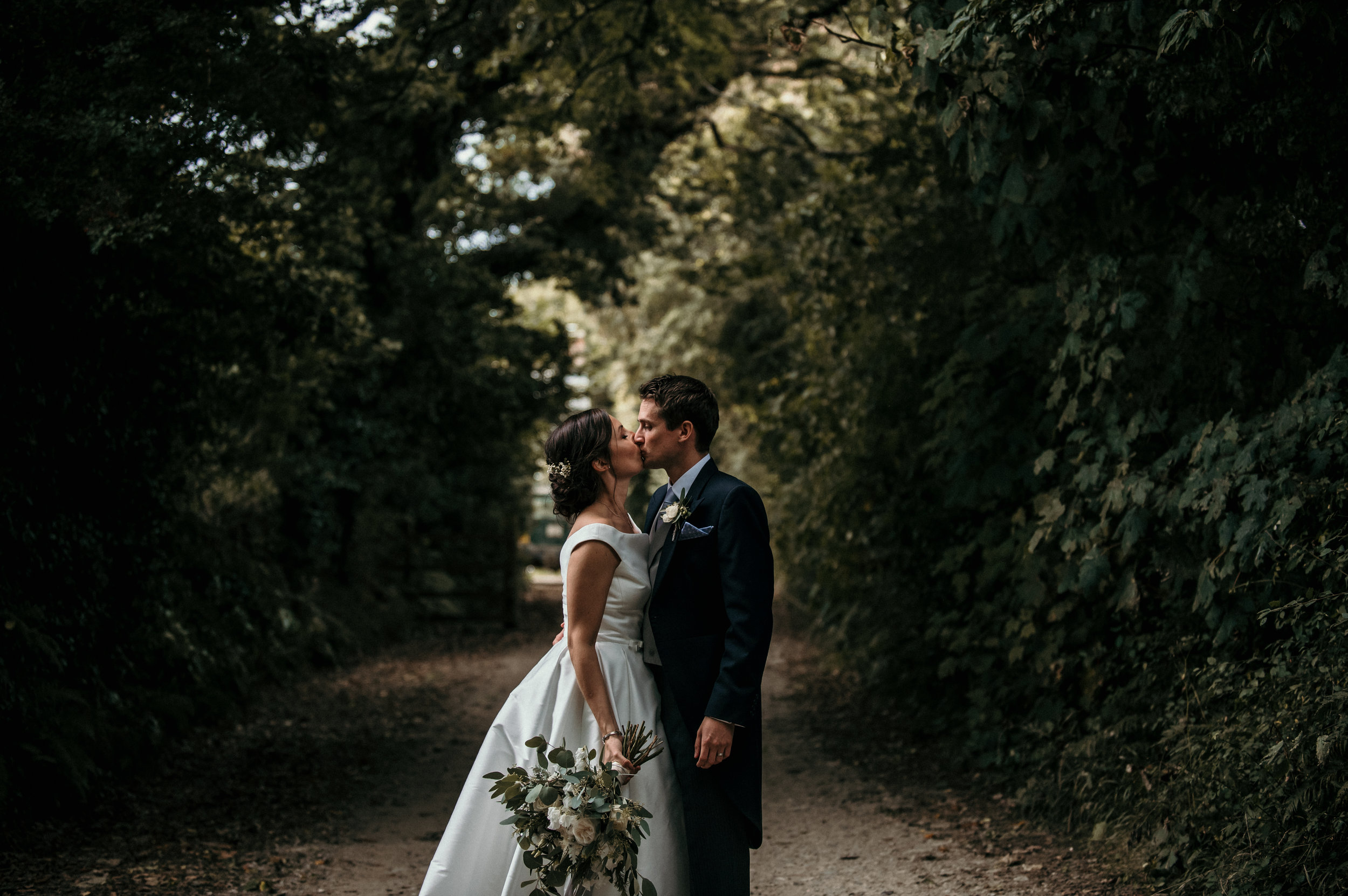 A bride and groom sharing a kiss in a wooded outdoor setting, the bride holding a bouquet of flowers, with green trees and natural light overhead.