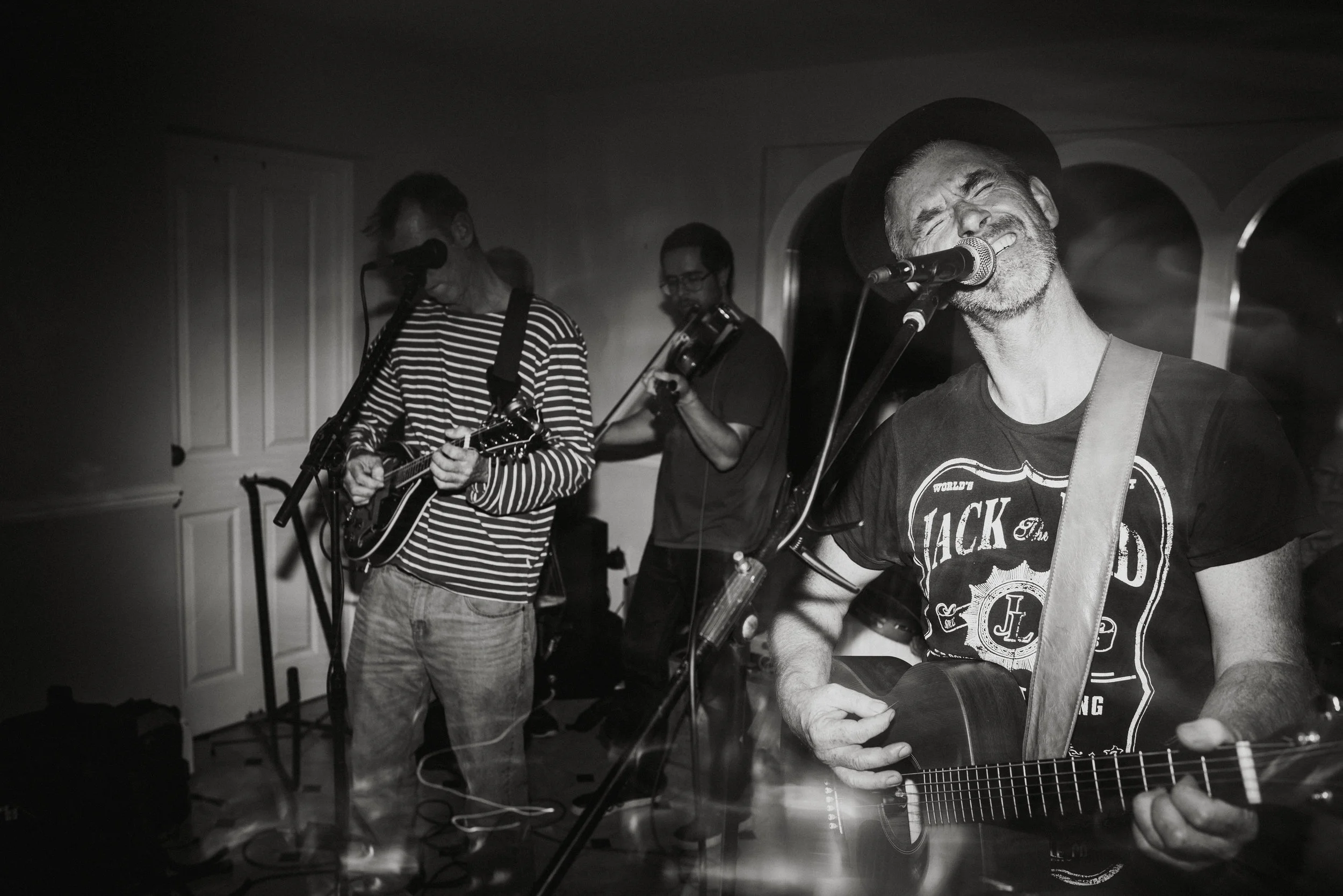 A black and white photo of a musical band performing, with a man singing and playing guitar in the foreground, a man with a violin in the middle, and a man with an electric guitar in the background.