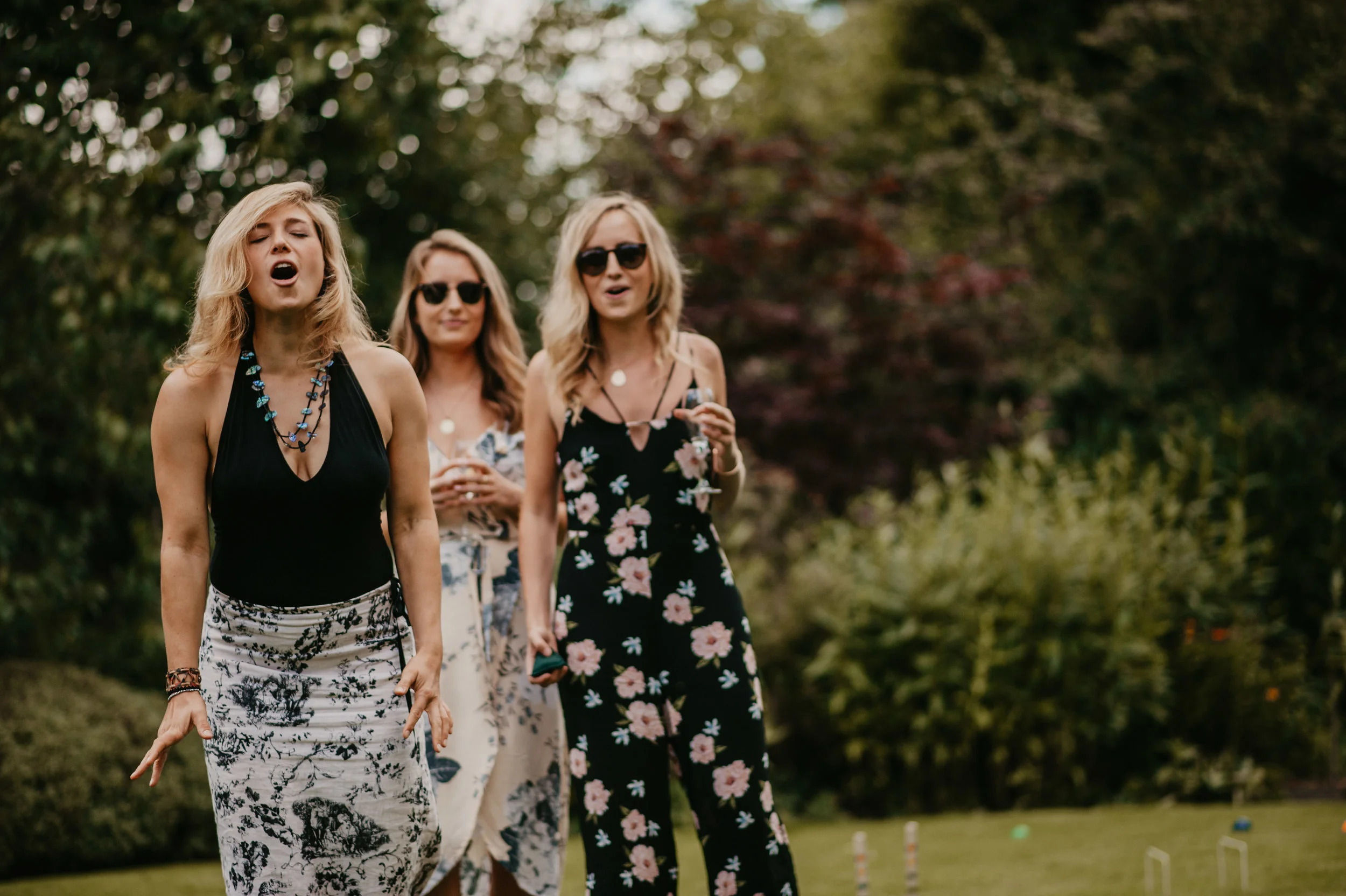 Group of three women outdoors, one singing with eyes closed, others holding drinks, dressed in floral and summery outfits, with lush greenery in background.