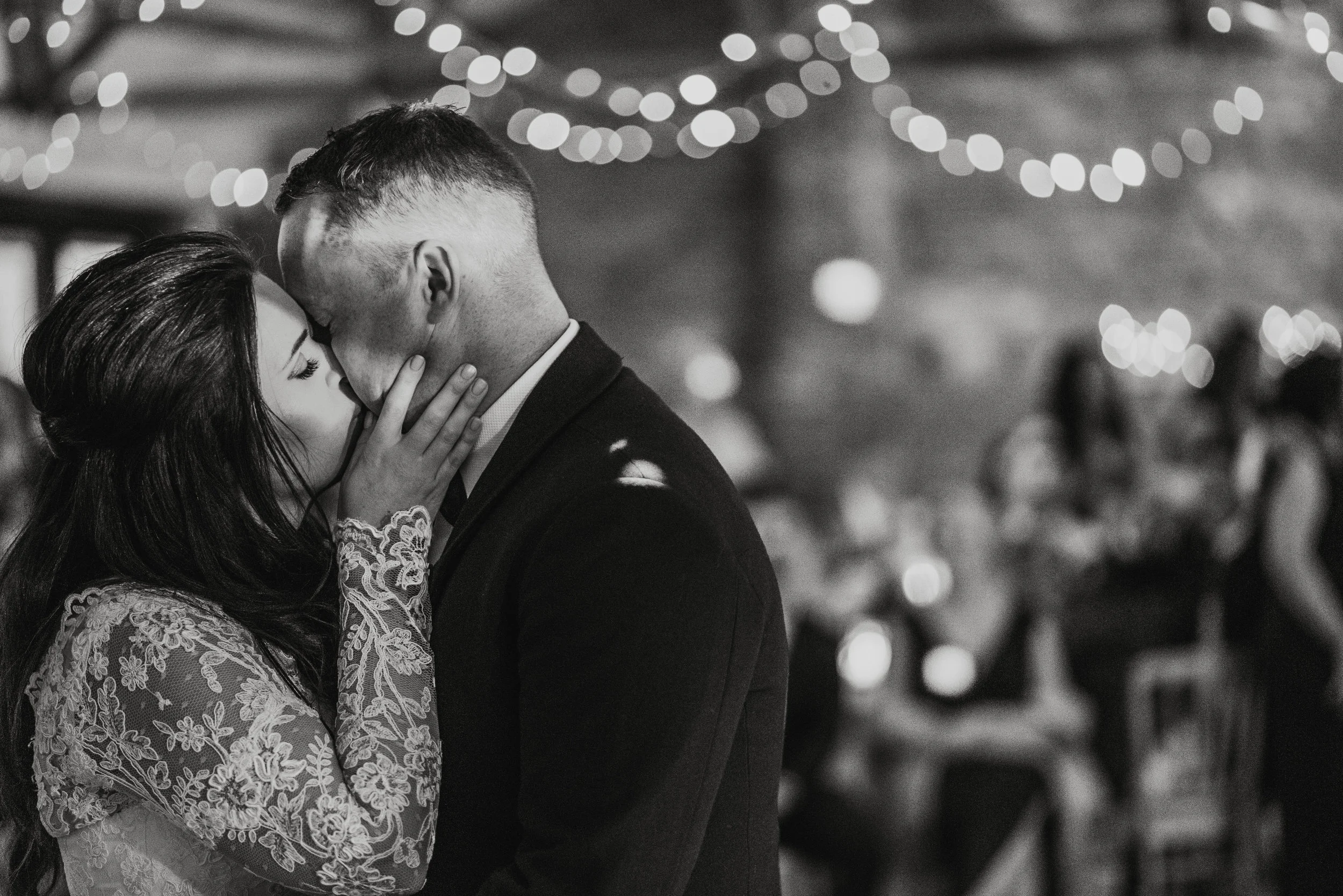 A black-and-white photo of a bride and groom sharing a kiss during their wedding reception, with blurred guests and string lights in the background.