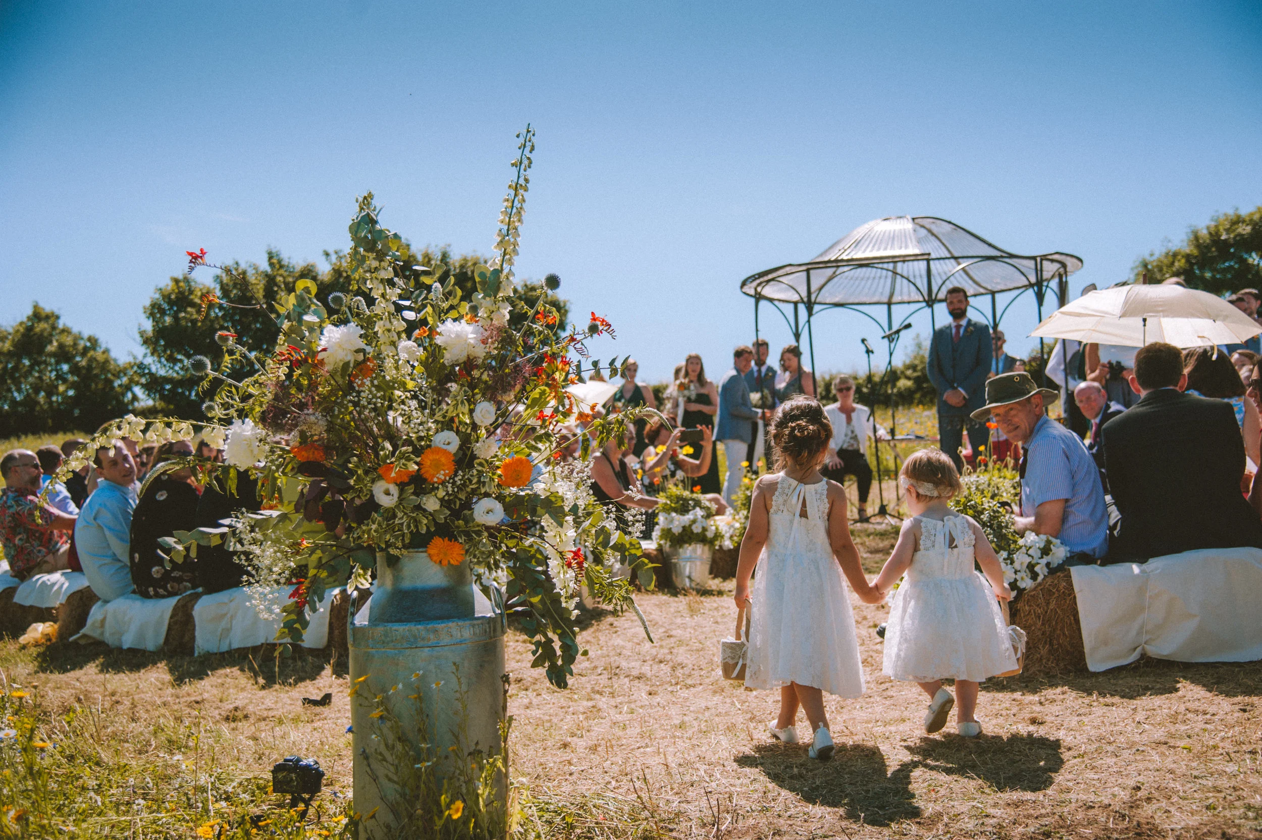 Two young girls in white dresses holding hands, walking towards an outdoor wedding ceremony. In the background, people seated on hay bales, a musician playing instruments, and a gazebo with a man in a suit standing underneath. A large floral arrangem