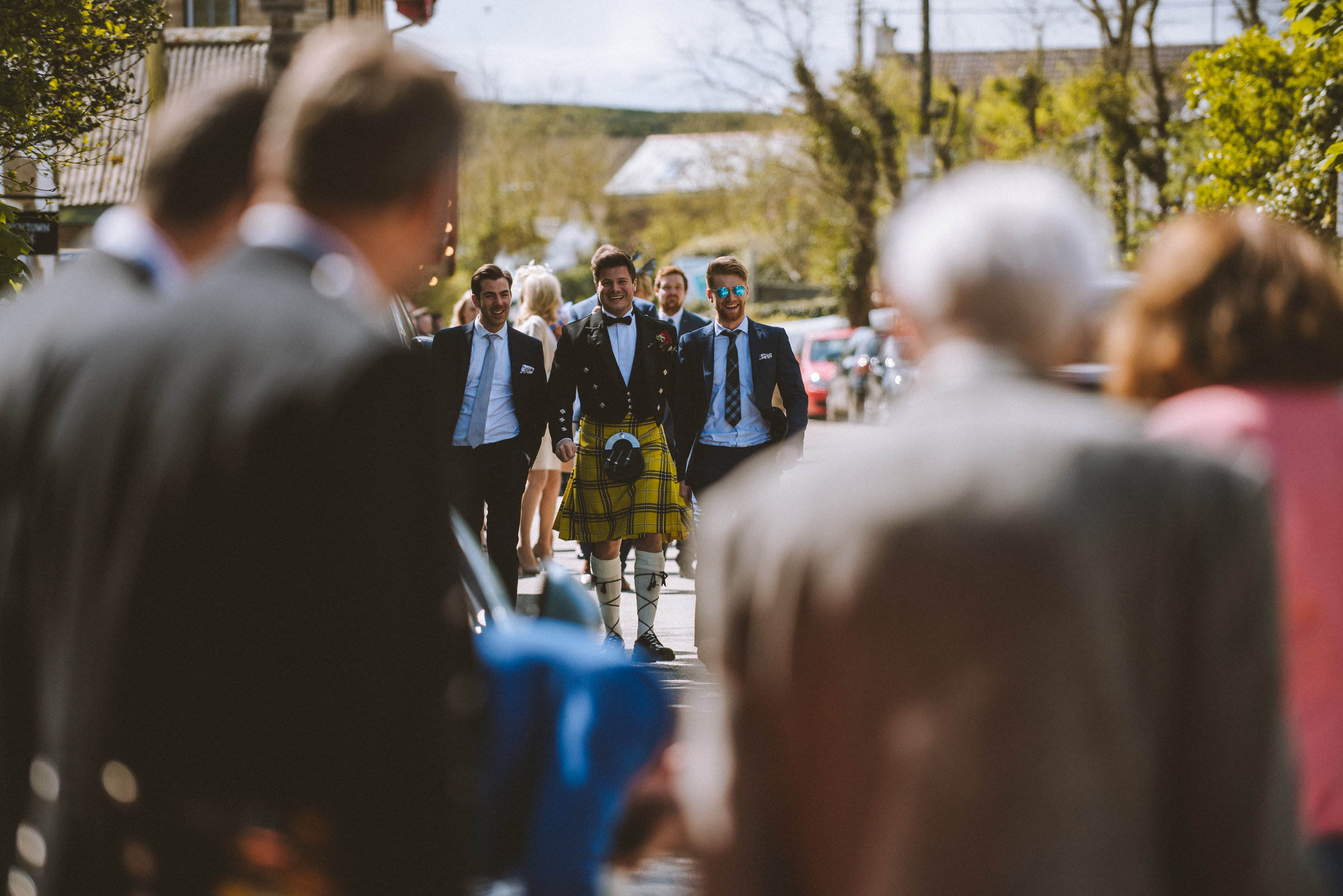 A man wearing a traditional Scottish kilt and formal jacket walking among a crowd of people, with several men in suits visible in the background outdoors on a sunny day.