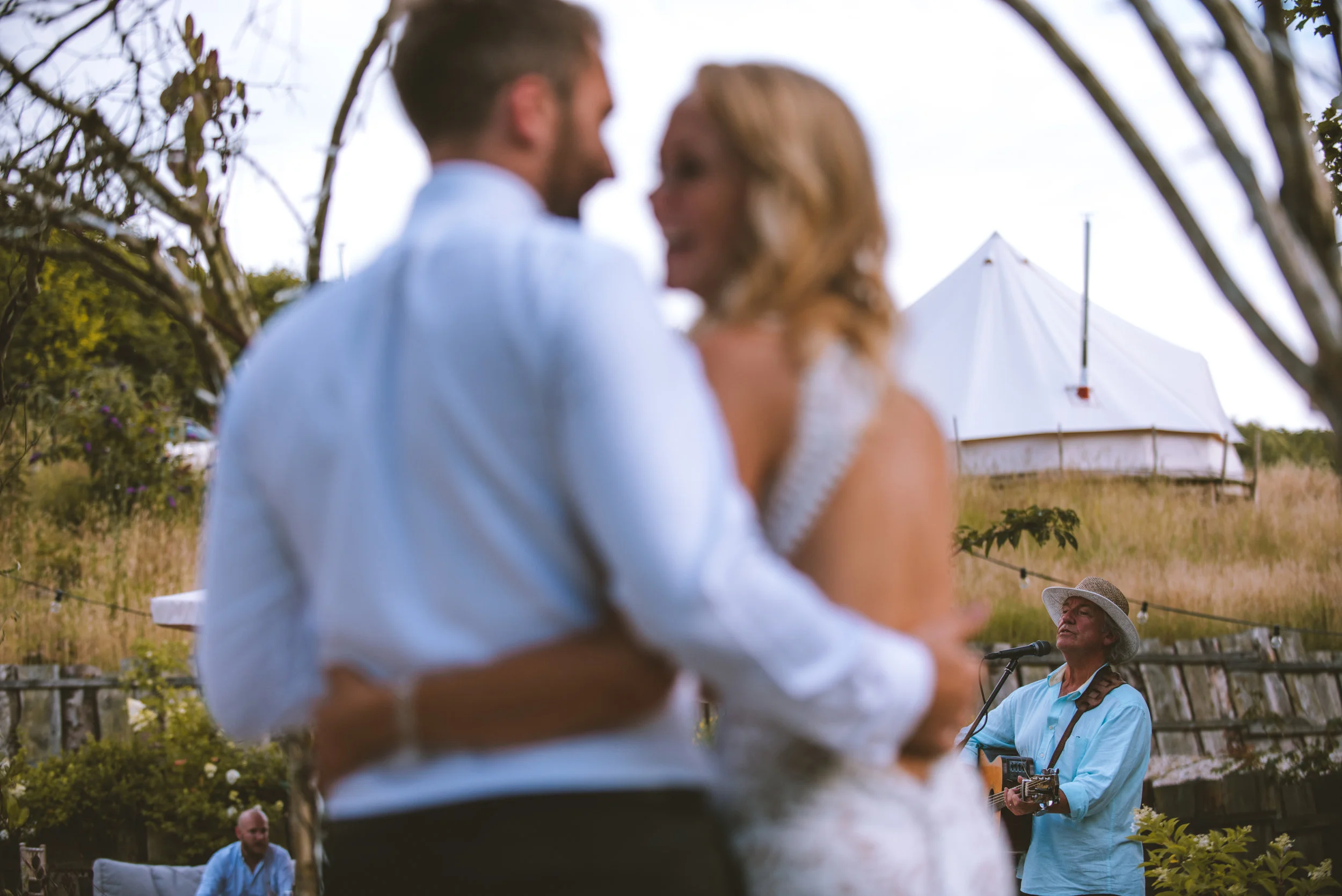 A couple dancing closely at an outdoor wedding ceremony, with a musician in the background playing guitar and singing into a microphone.