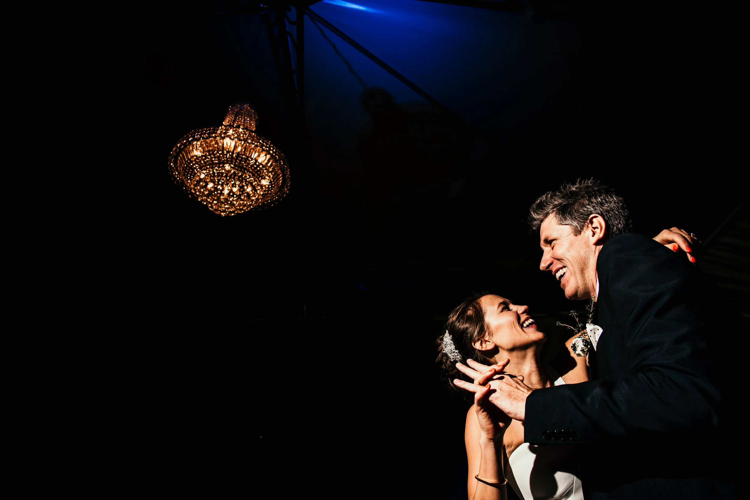 A bride and groom dancing and smiling at their wedding reception, with a chandelier overhead and a dark background.